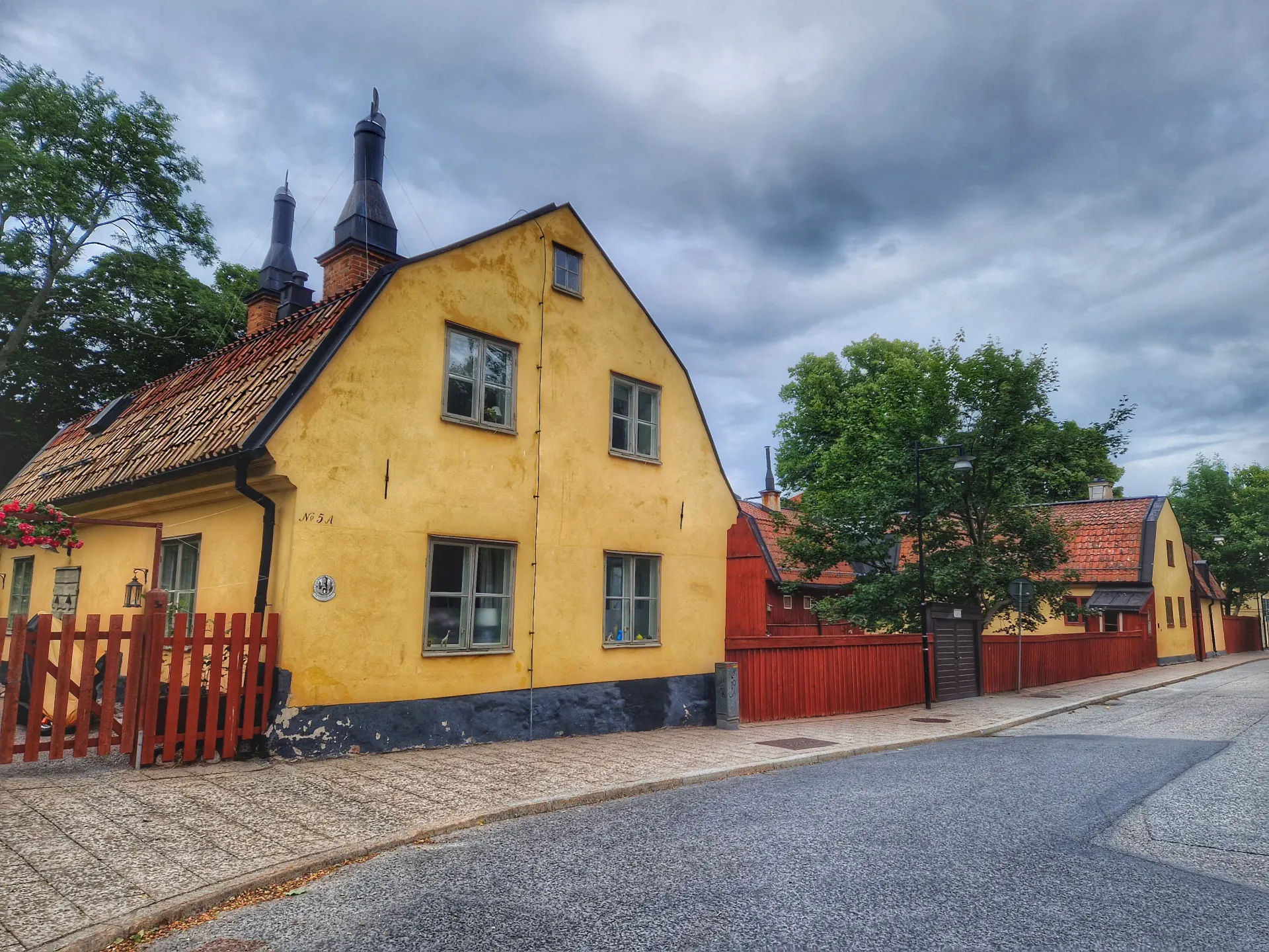 Gul byggnad med rött staket längs kullerstensgata. Molnig himmel