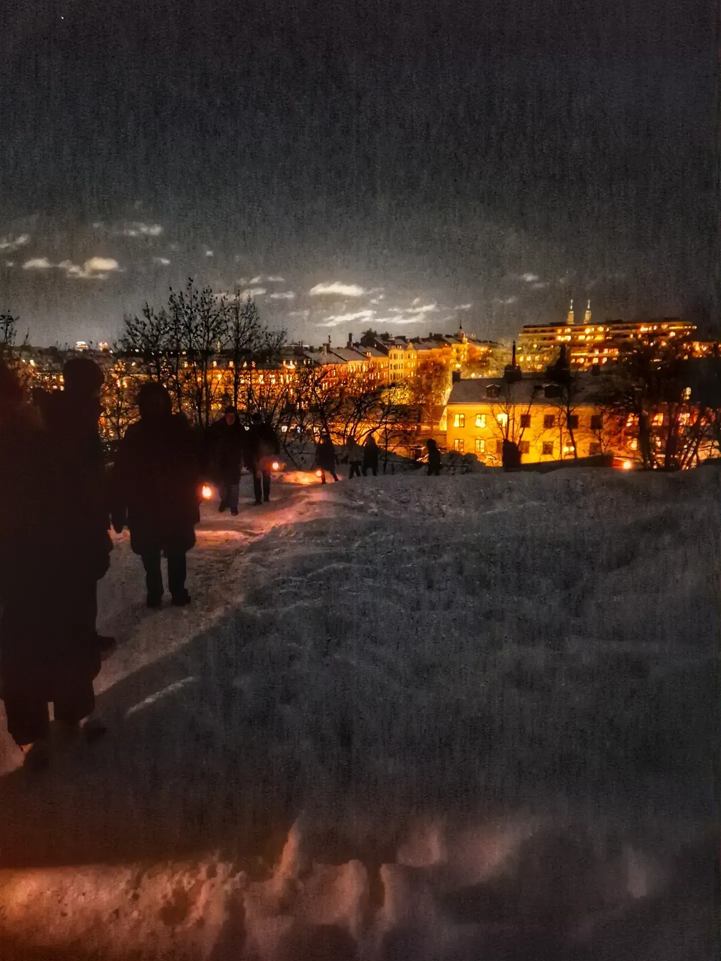 Människor med lyktor går på snöig stig. Stadens ljus i bakgrunden. Natt