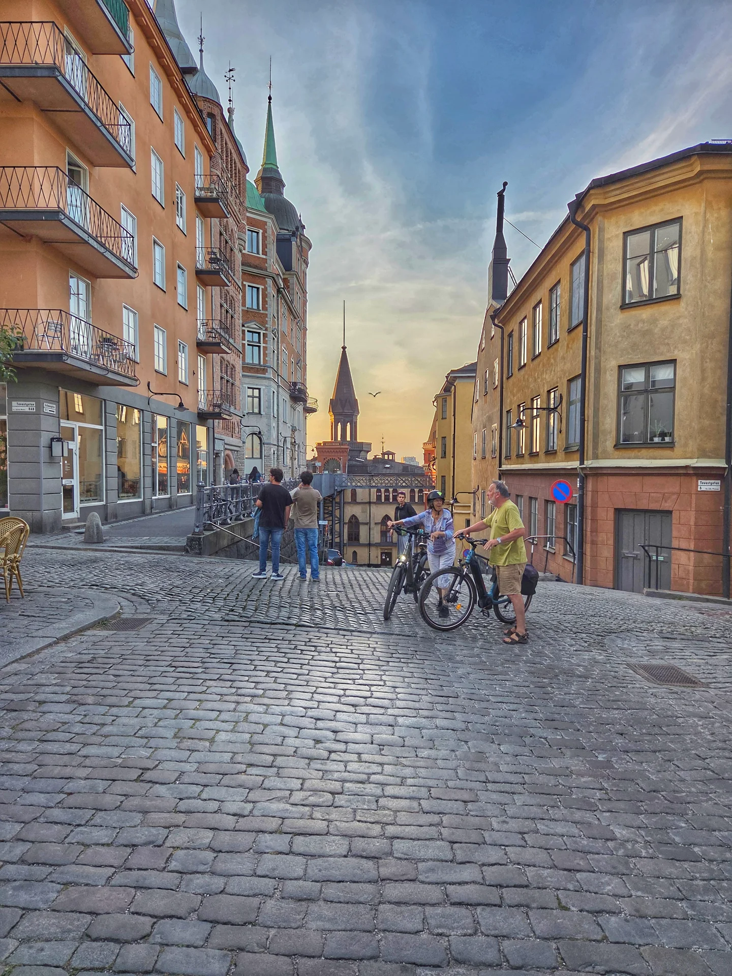 Stadsvandring Mariaberget: Kullerstensgata med historiska hus, människor med cyklar och torn i kvällssol