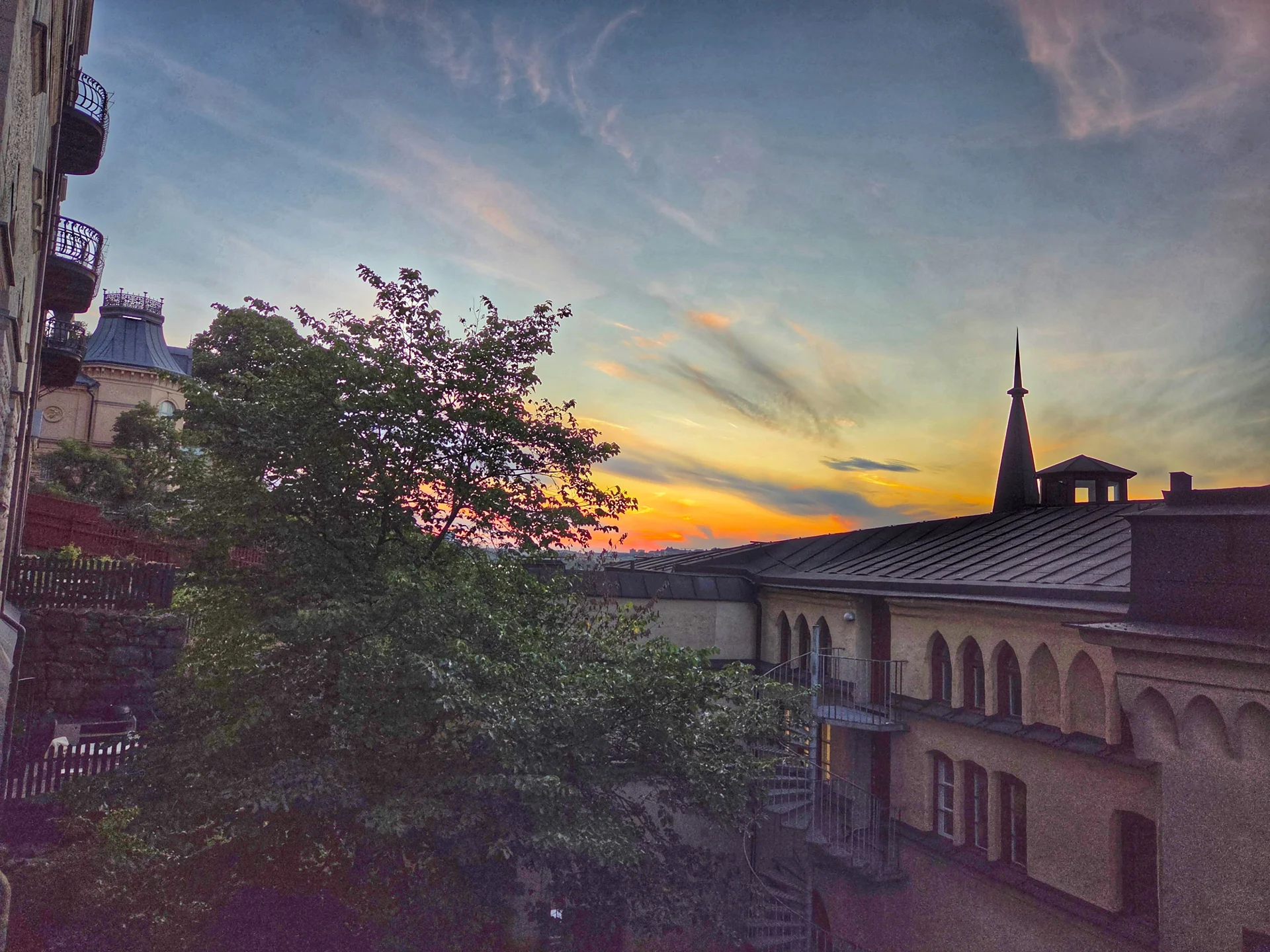 En vy från en stadsvandring på Mariaberget i solnedgång. Gamla hus, träd och en spira mot färgglad himmel