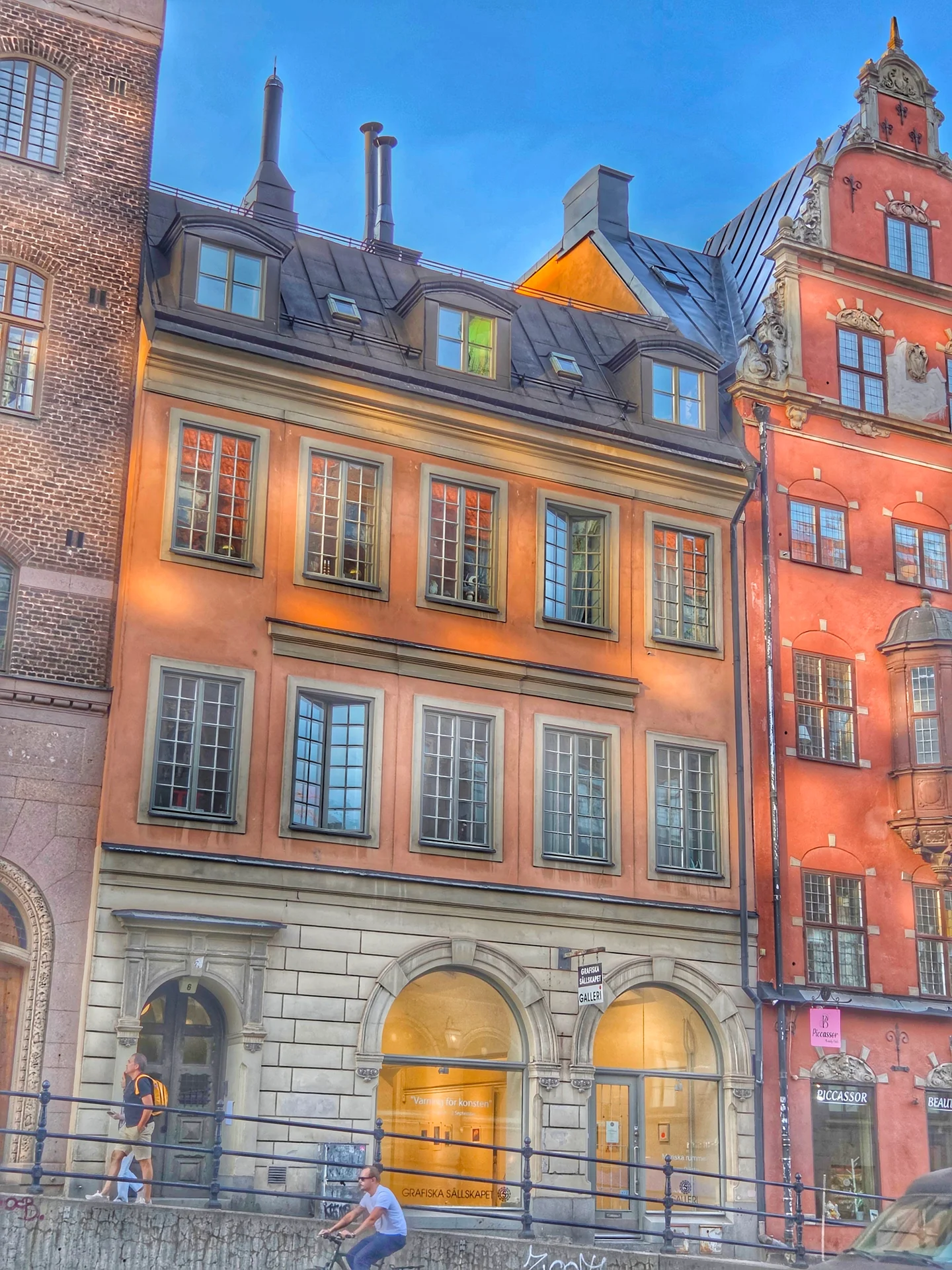 Solbelysta fasader på historiska stadshus. En man går, en cyklar på gatan framför dem