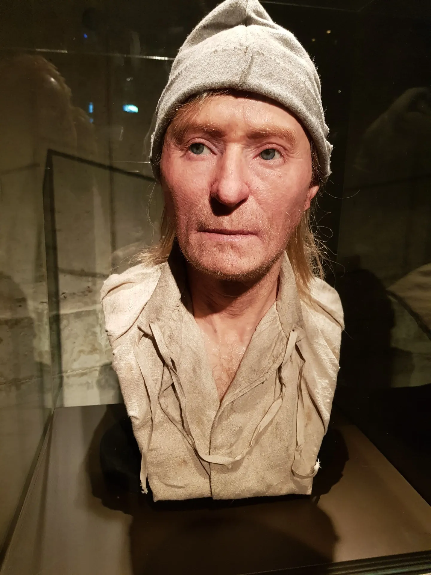 Byst av man med mössa och ljus skjorta. Står i en glasmonter