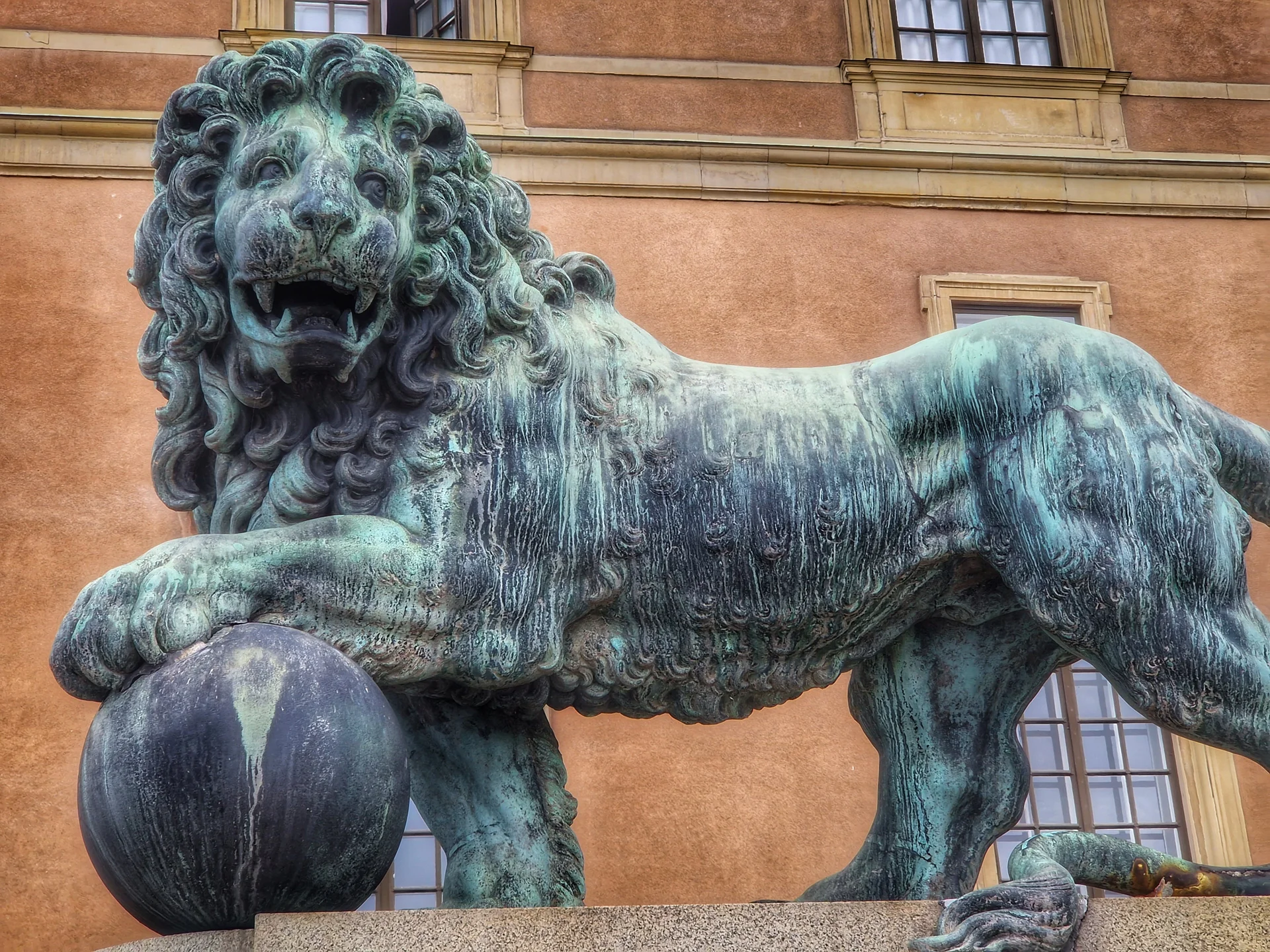 Grönpatinerad bronslejonstaty med tass på kula framför fasad. Passar för Stadsvandring Gamla Stan