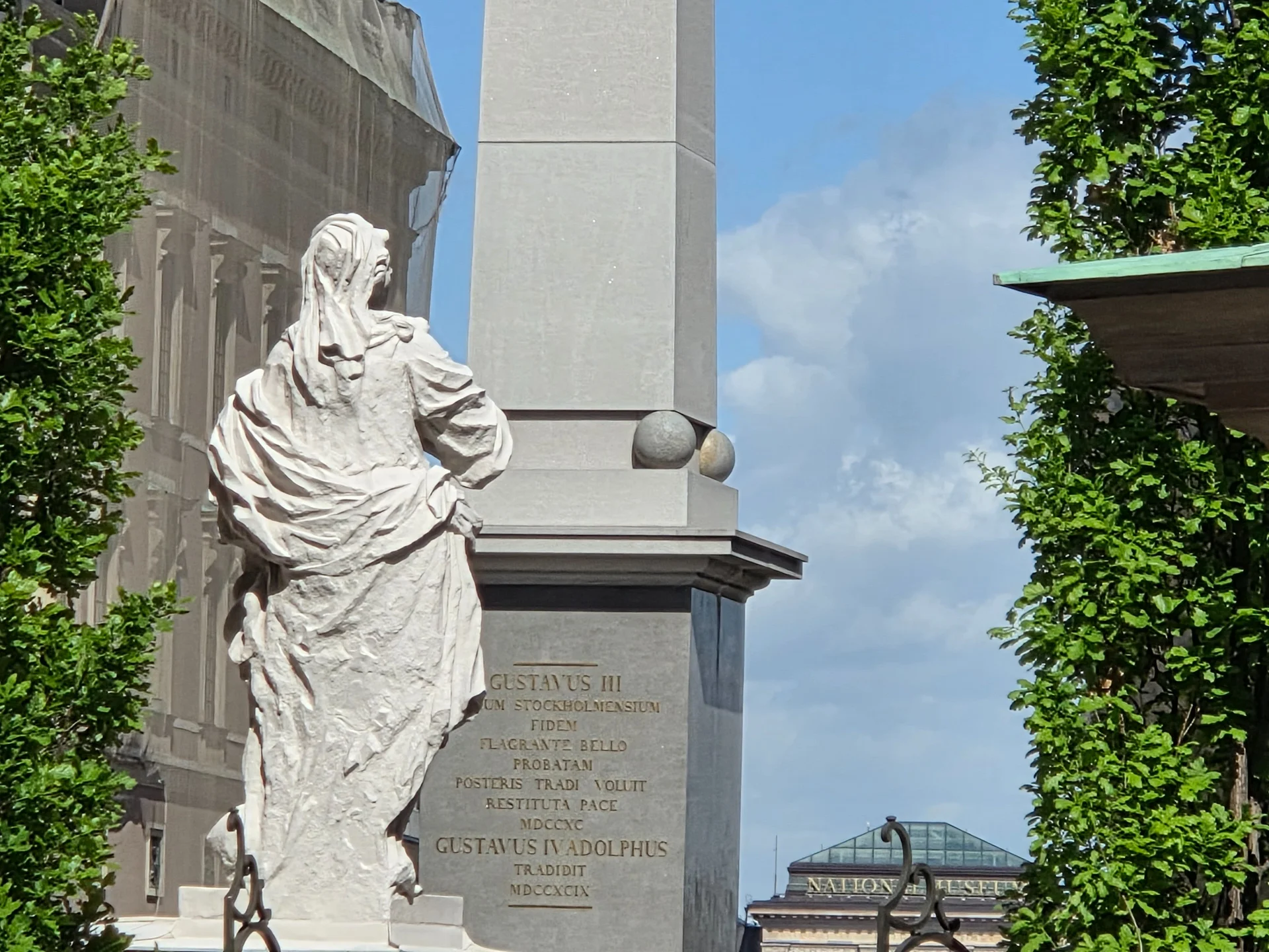 Vit staty, obelisk med text om Gustav III. Gröna träd och Nationalmuseum i bakgrunden. Stadsvandring Gamla Stan
