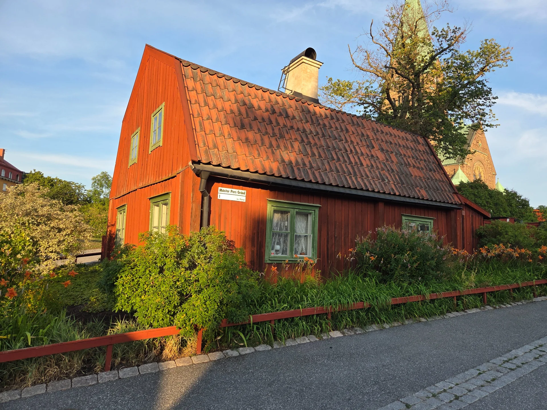 Rött trähus på Mäster Pers Gränd med gröna fönster och buskar. Kyrktorn syns i bakgrunden