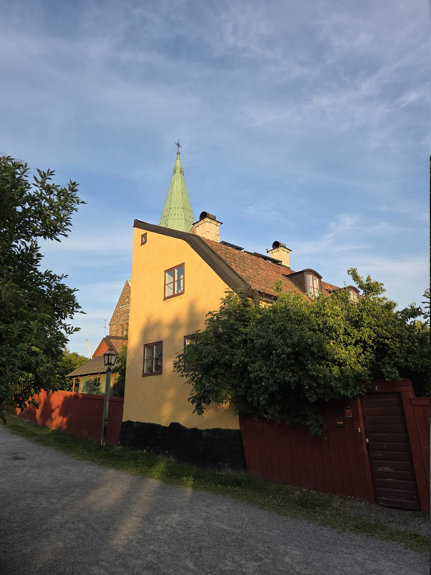 Gult hus med tegeltak och grön kyrkspira mot blå himmel. Grusväg, rött staket och lummig grönska i förgrunden