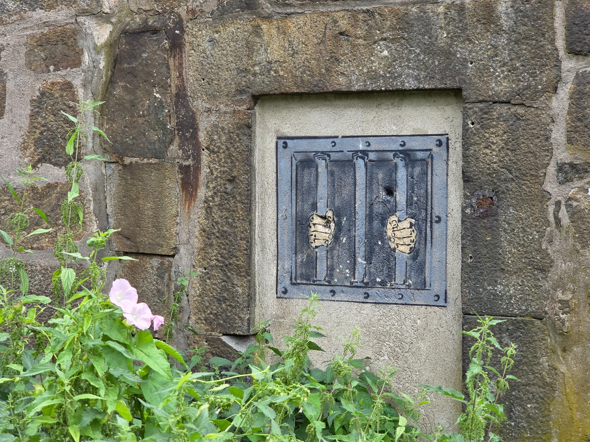 Konstinstallation med fängelsegaller och händer på en stenmur, med gröna växter och rosa blommor. Långholmen fängelse
