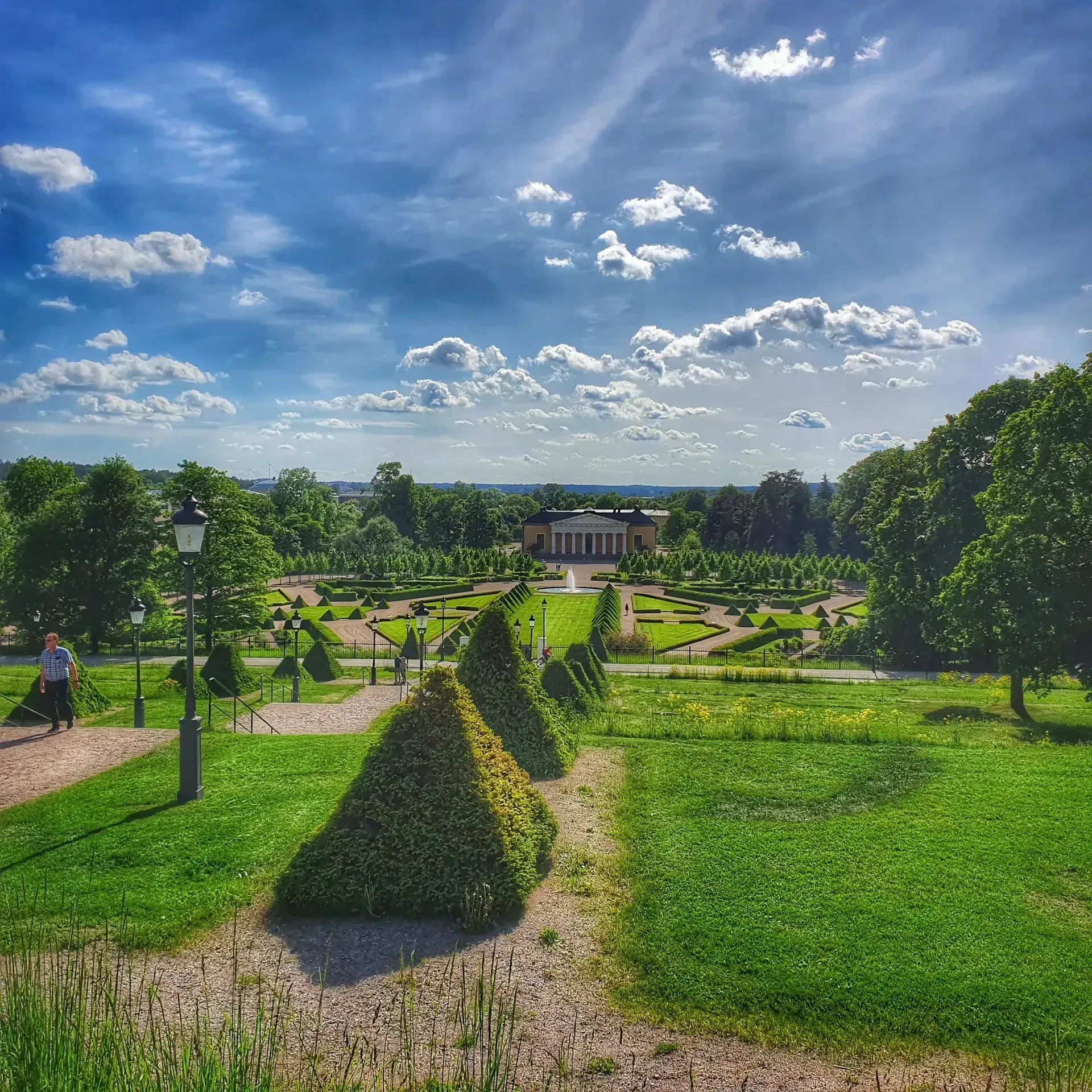 Park med en lång, formell trädgård och byggnad i fonden. Blå himmel med moln. Stadsvandring Uppsala