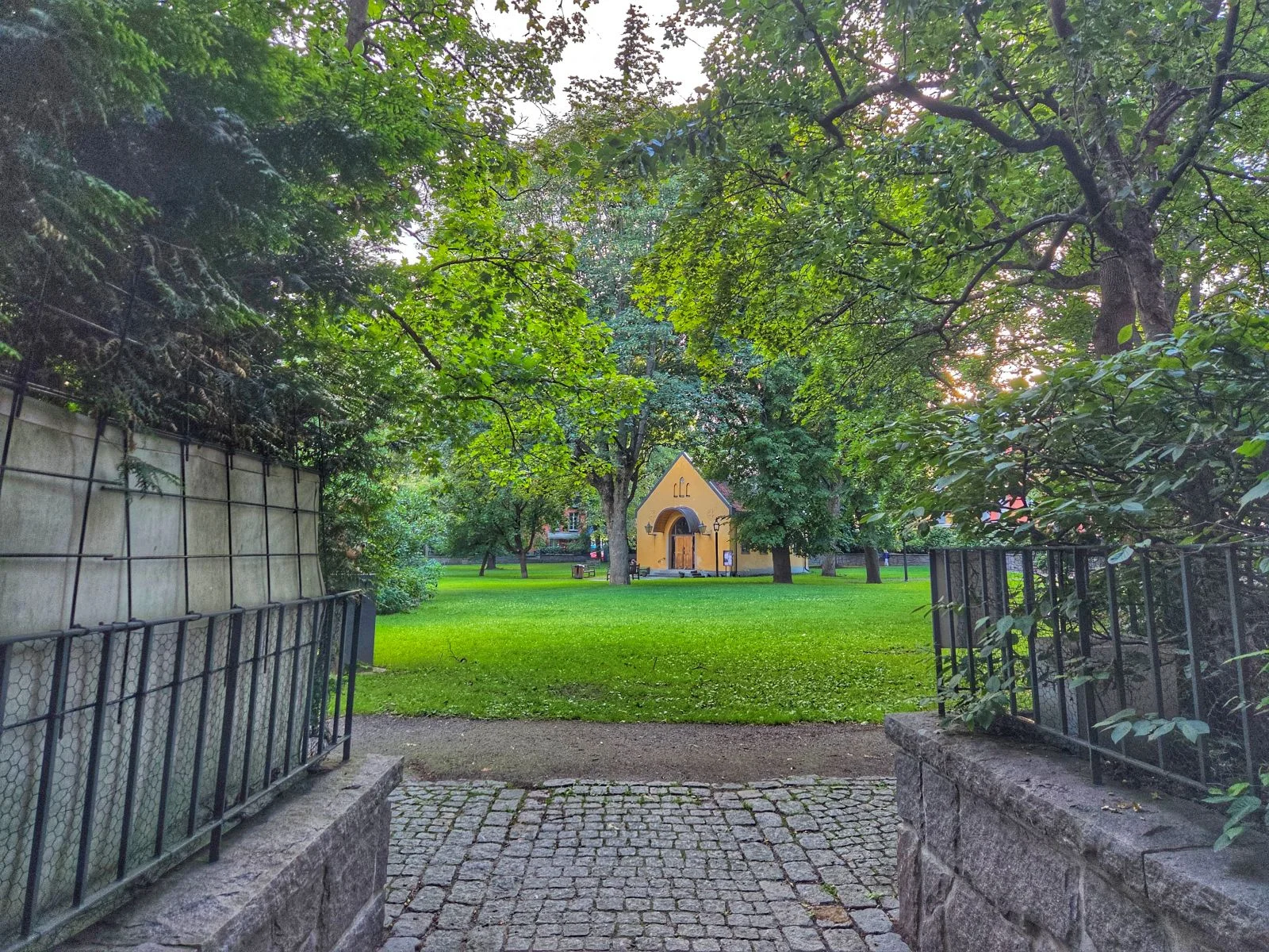 Kullerstensgång leder mot en gul byggnad i en grön park, omgiven av träd och staket