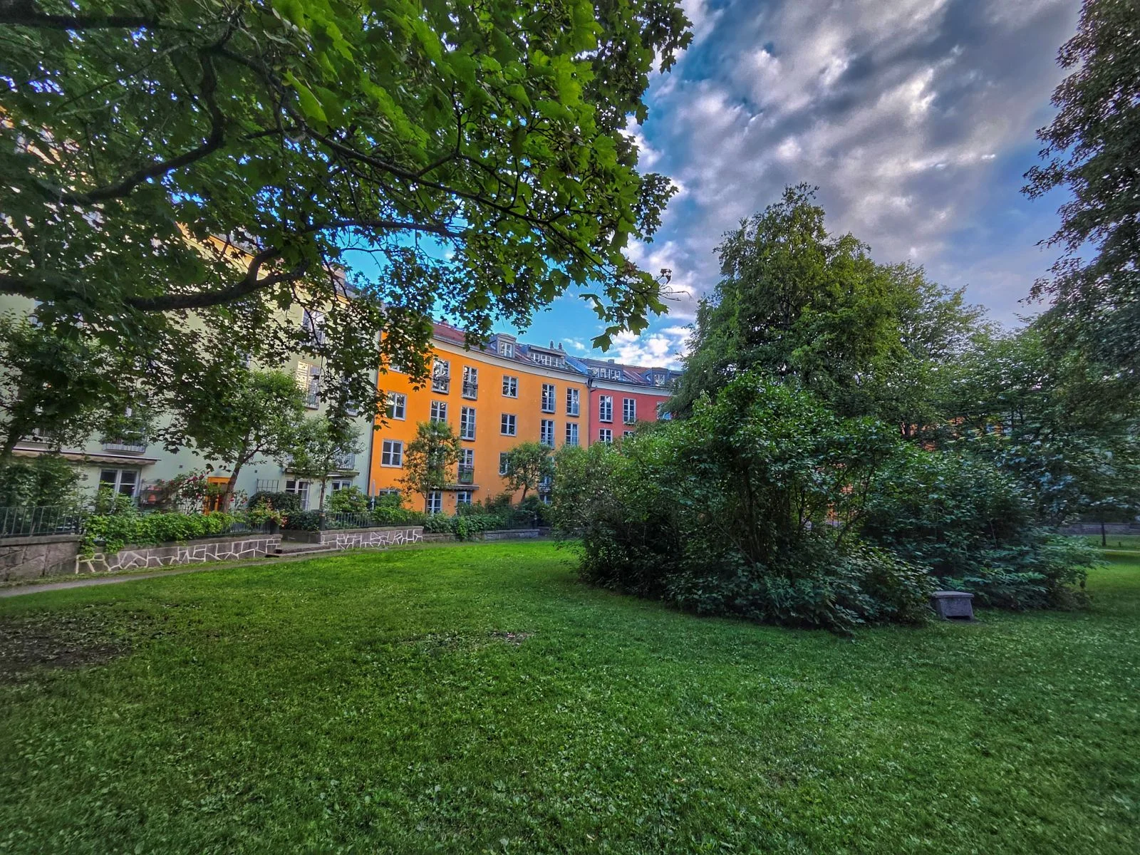 Grön gräsmatta framför färgglada flerbostadshus med lummiga träd och buskar under en molnig himmel