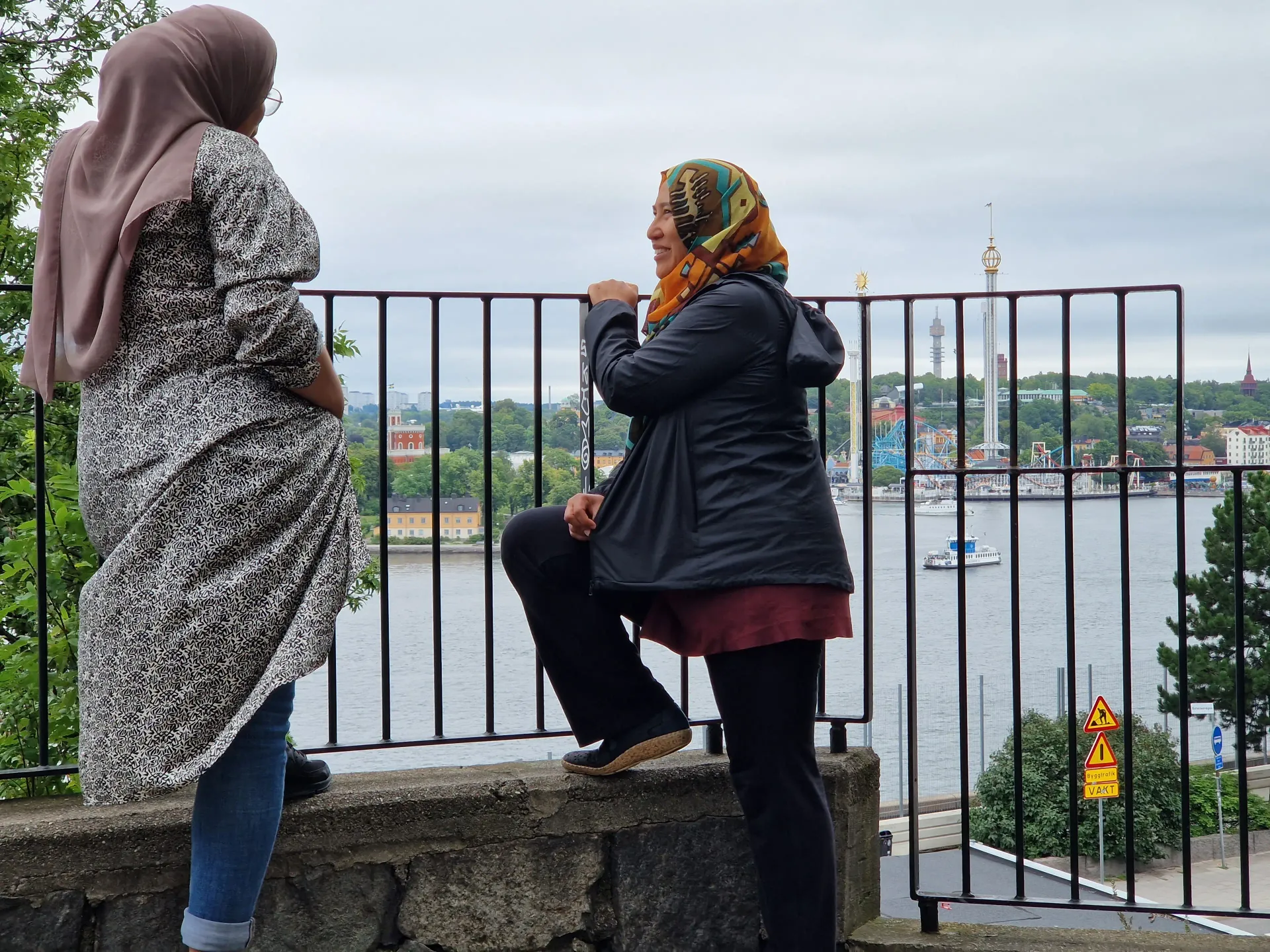 Två kvinnor står vid ett räcke med utsikt över vatten och byggnader. En kvinna bär hijab. Vy från Stadsvandring Södermalm