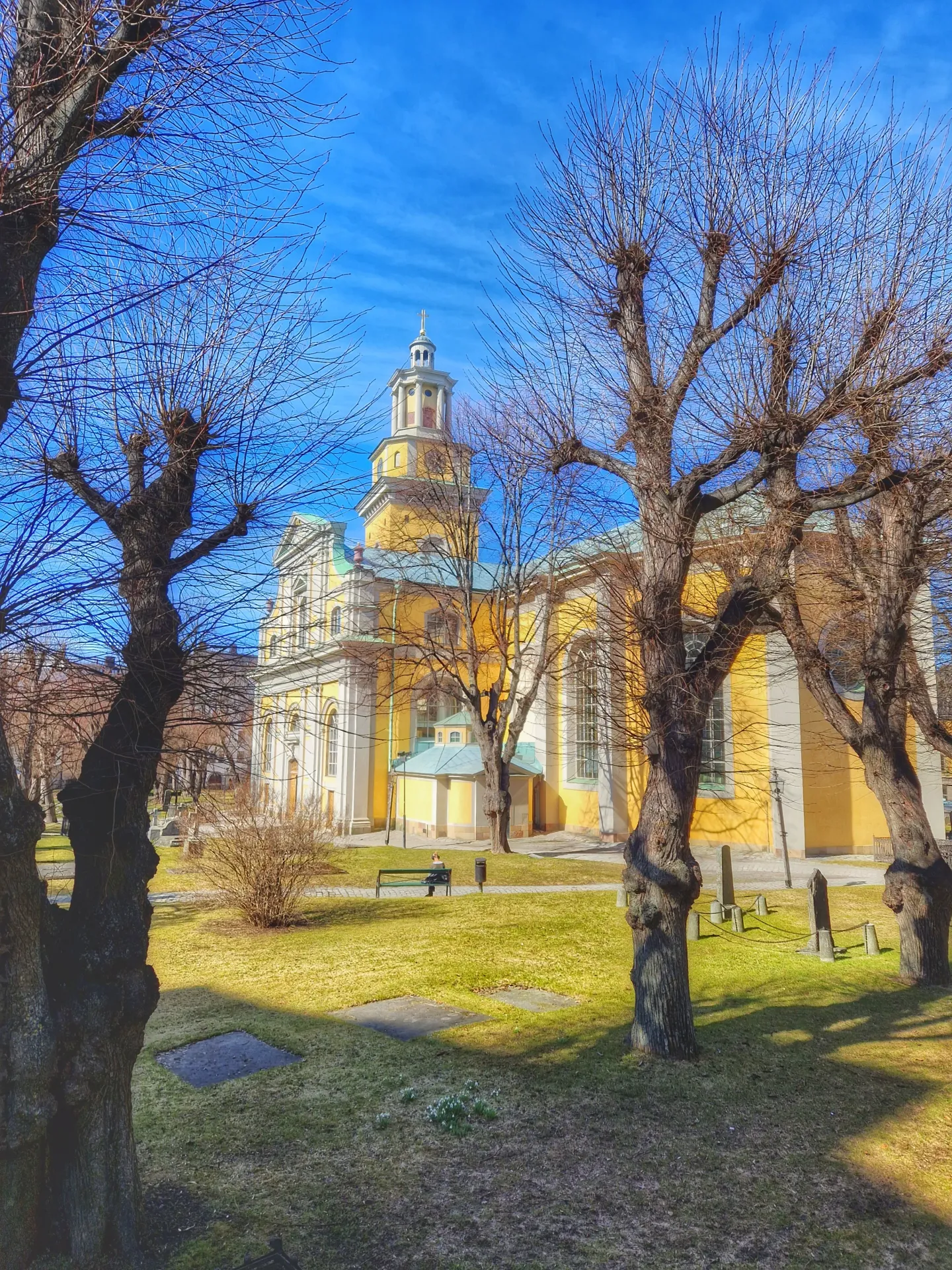 Gul kyrka med torn, träd utan löv, gräsmatta och blå himmel. Person på bänk. Gravstenar syns också