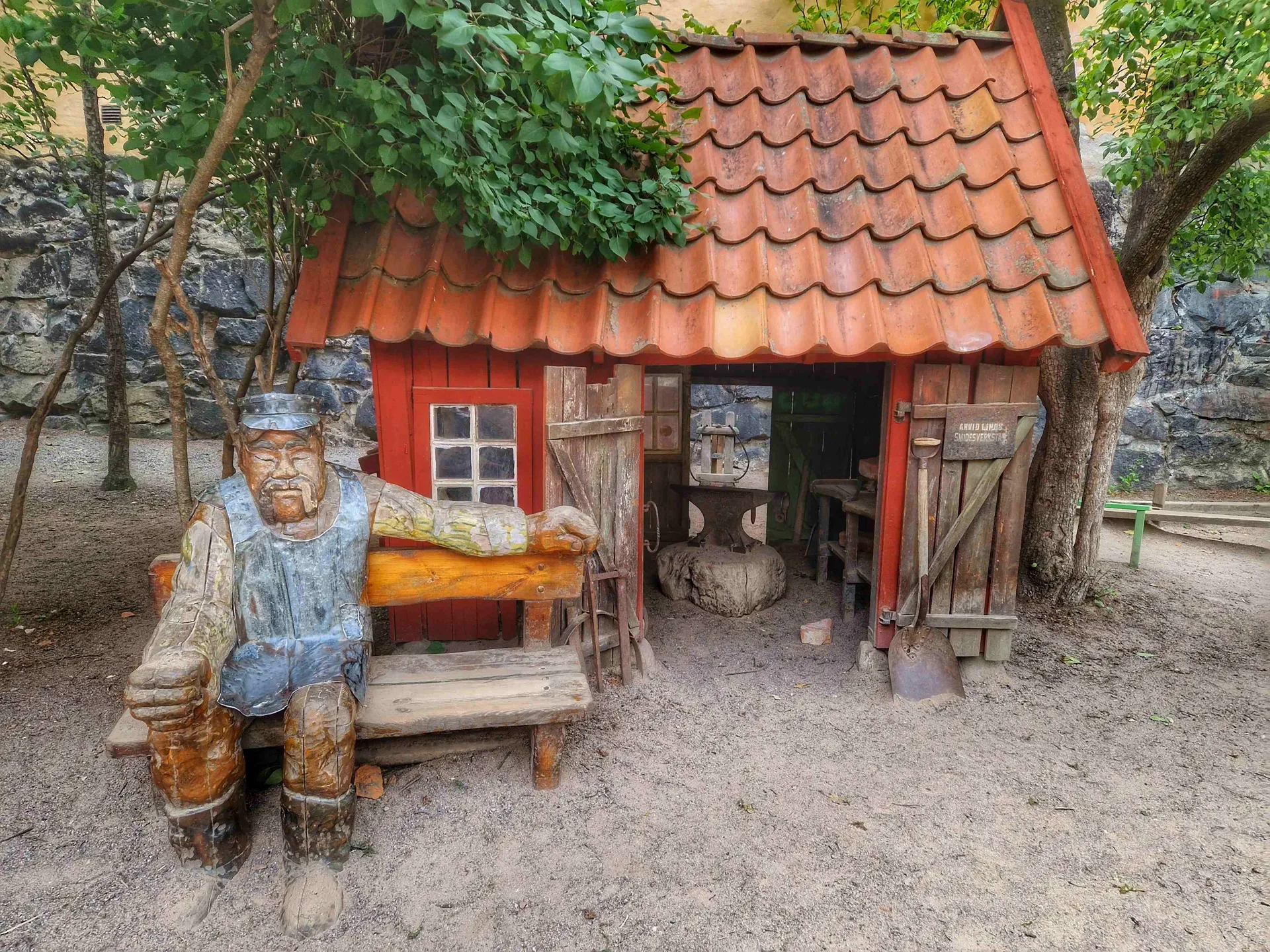 Liten smedja med rött tegeltak och trädörrar. En träskulptur sitter på bänk framför