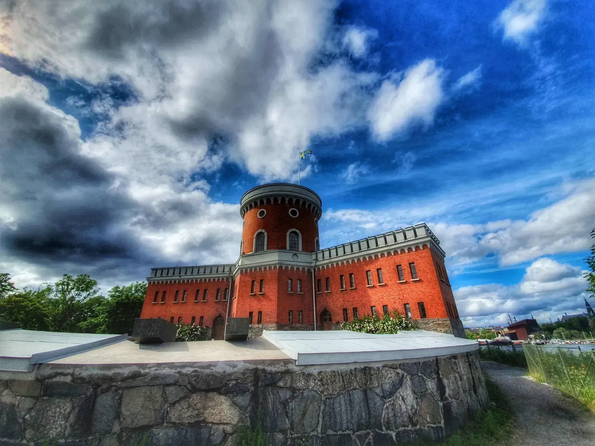 Kastellholmen, Ö i Stockholm: Röd tegelbyggnad med torn under blå himmel med vita moln. Svenska flaggan vajar. Mur i förgrunden