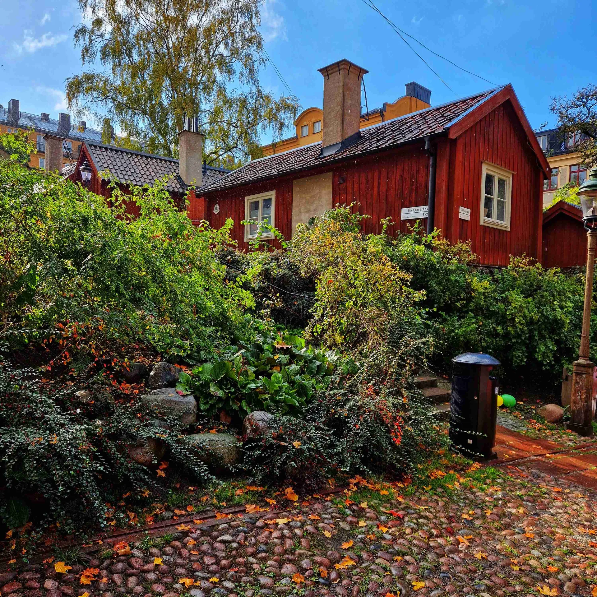 Rött trähus vid kullerstensgata, omgivet av grönska. Skylt med texten Skeppergränd. Stadsvandring Fogelström
