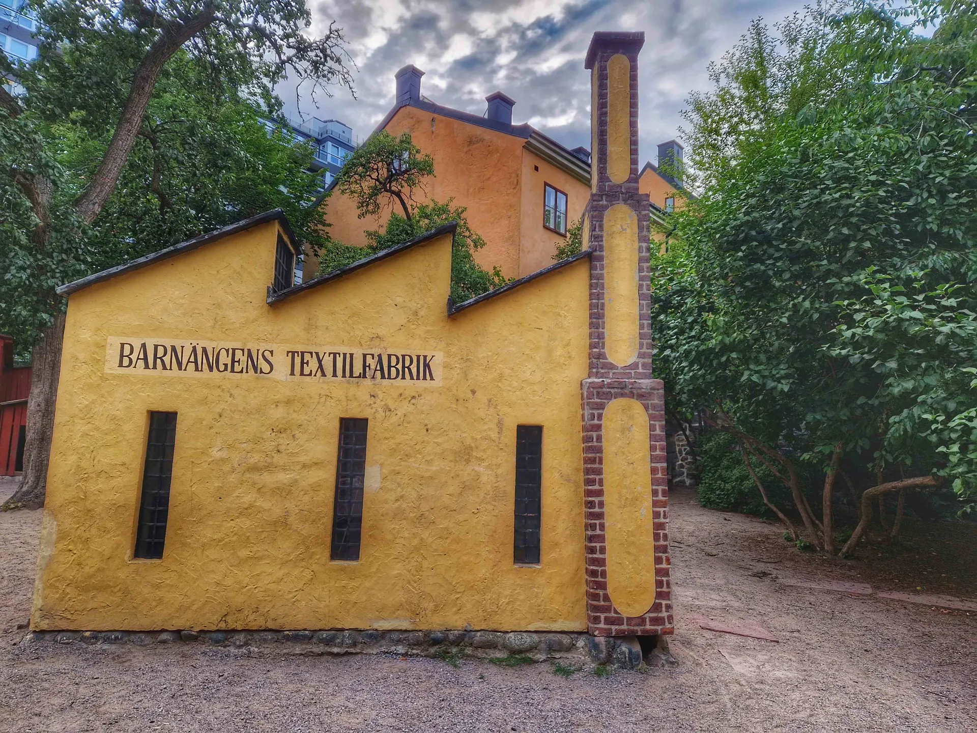 Gul byggnad med texten "Barnängens textilfabrik". En skorsten av tegelsten syns till höger
