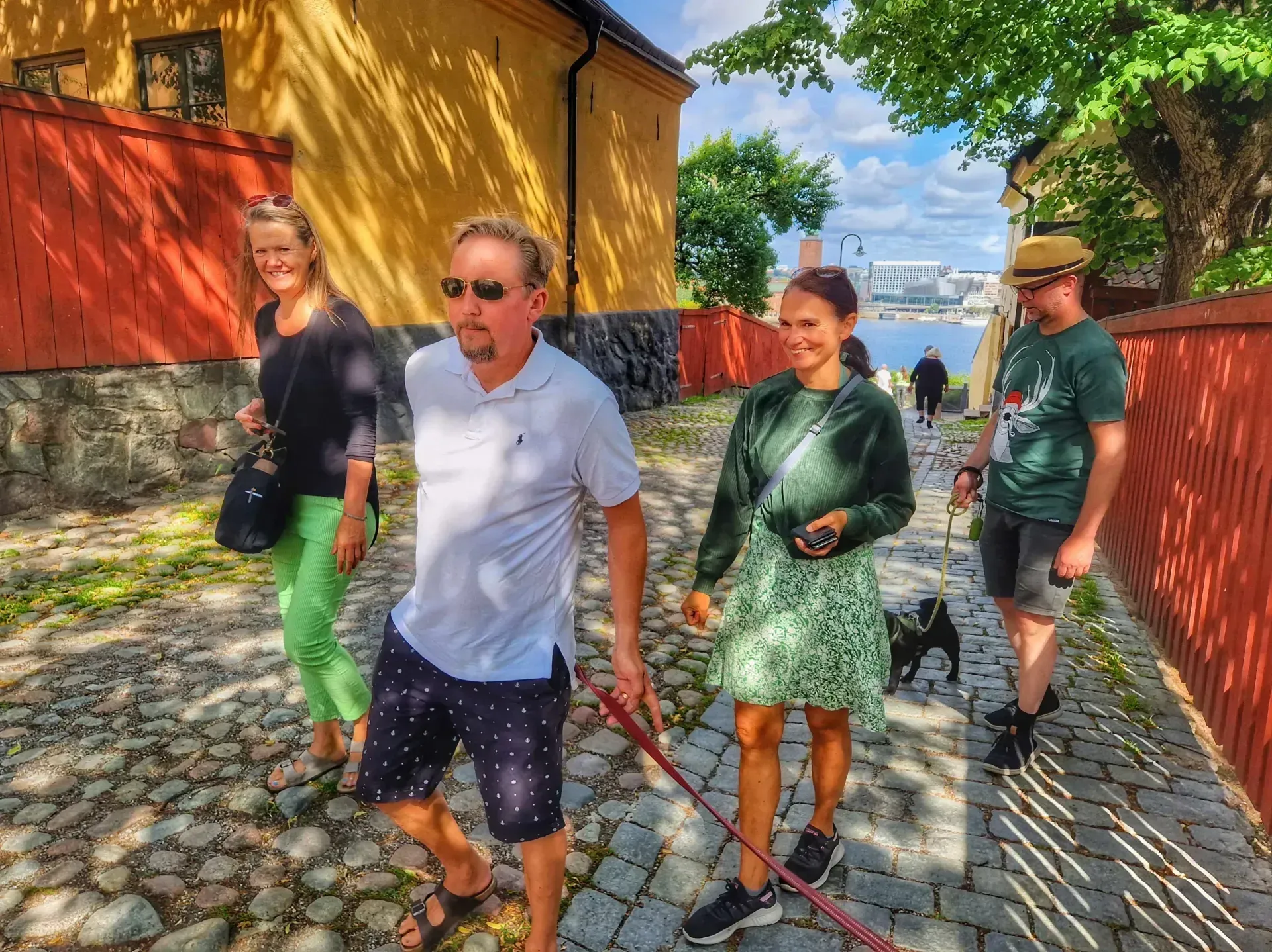 Fyra personer och en hund promenerar på kullersten. Gul byggnad och rött staket syns