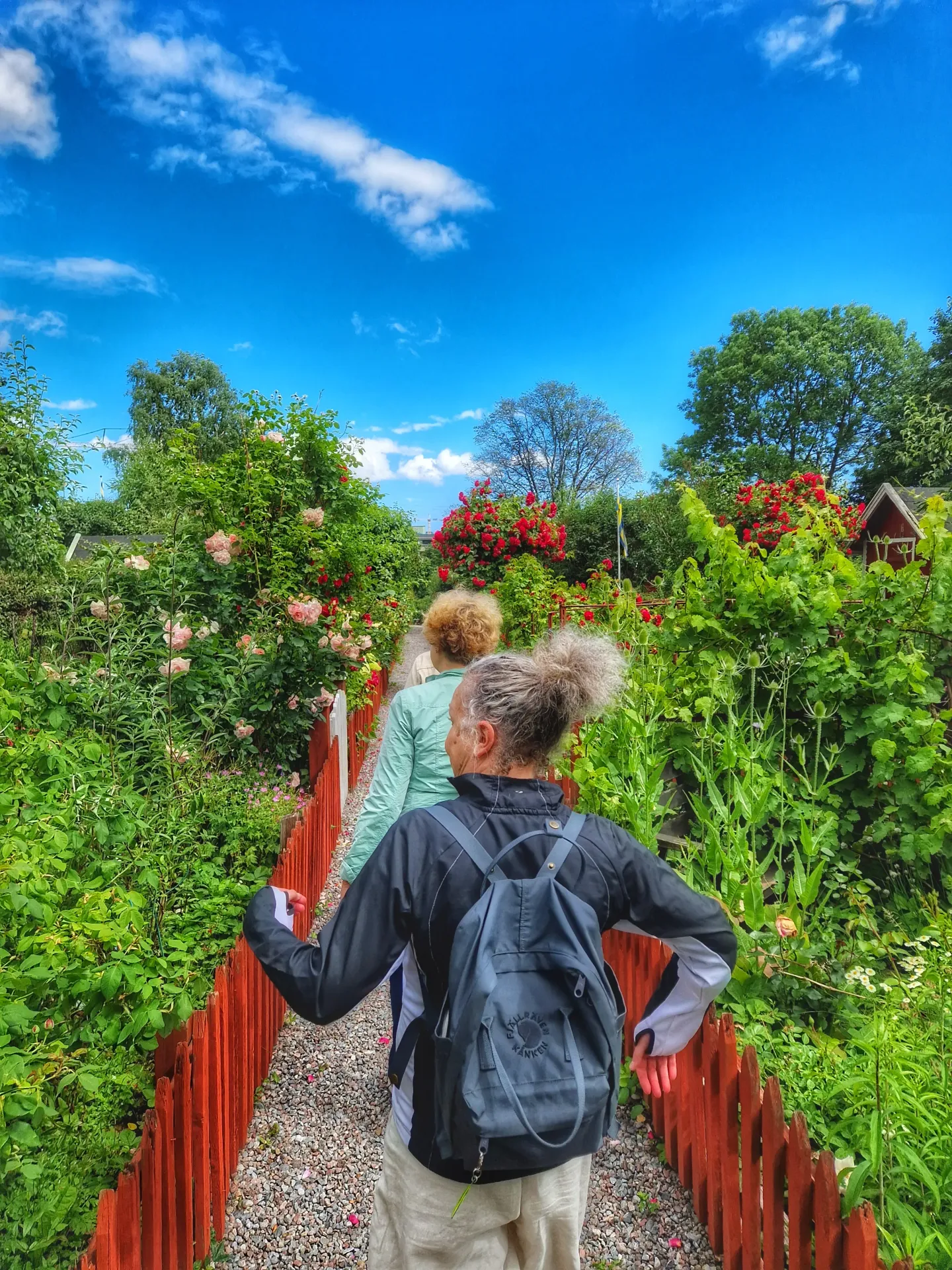 Två personer går längs en grusgång med rött staket och blommor. Blå himmel