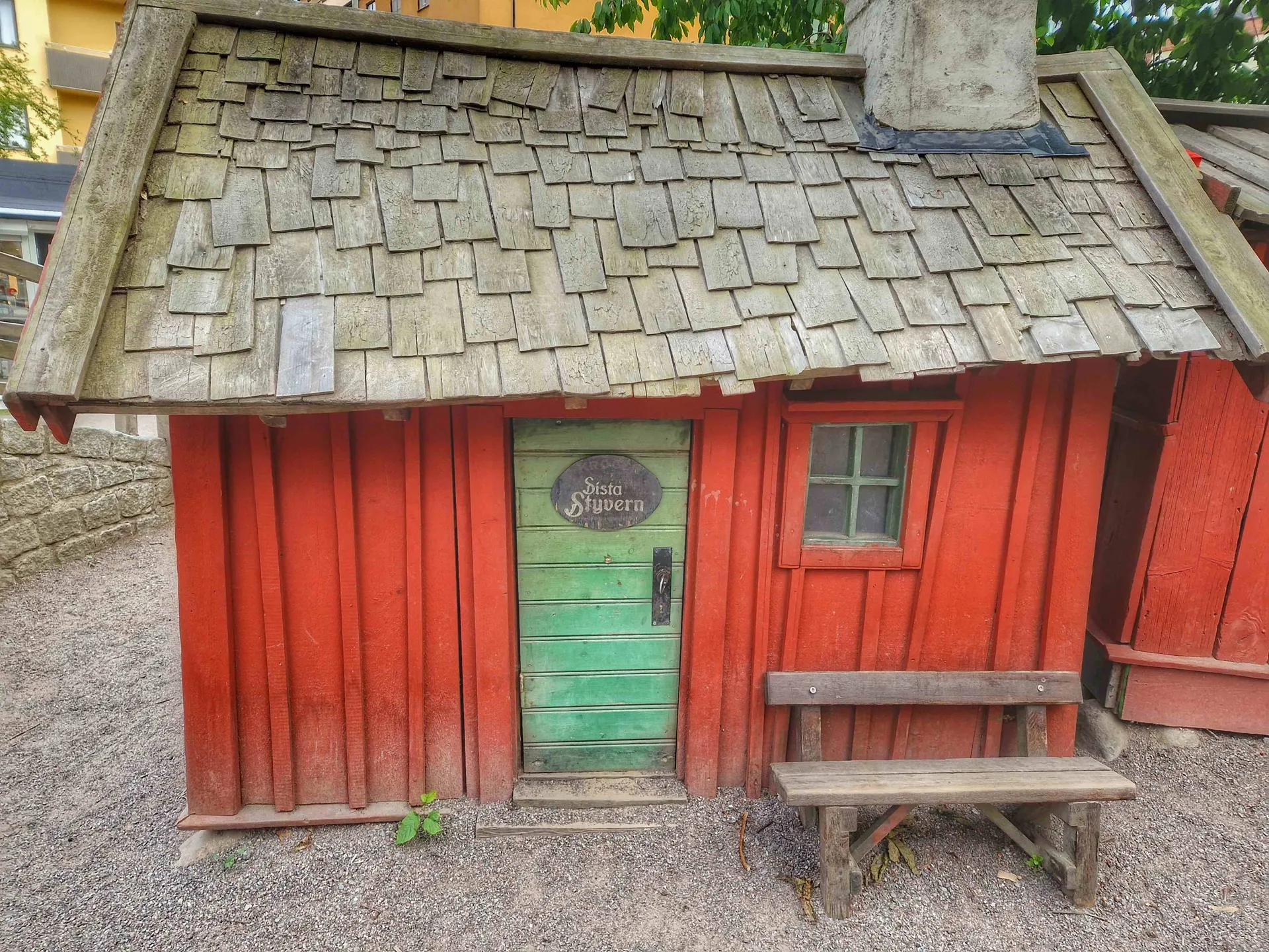Rött trähus med grön dörr och spåntak. Bänk framför. Skylt "Sista Styvern"
