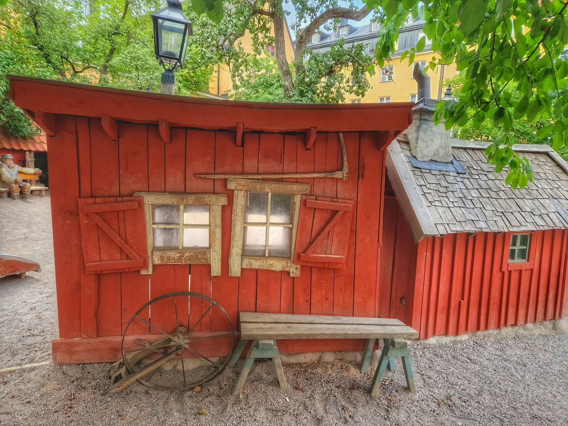 Röd timmerstuga med fönsterluckor, trähjul och bänk. Tegeltak på intilliggande byggnad