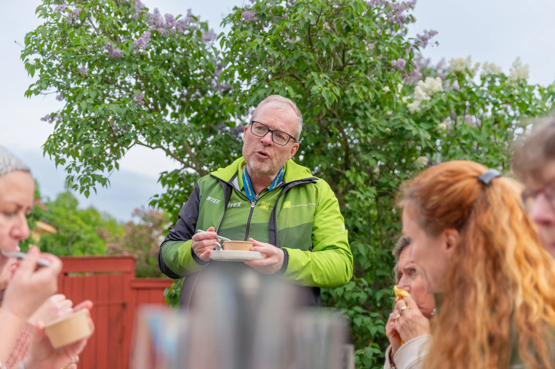 Person med skål och sked i handen pratar med en grupp. Syrener i bakgrunden