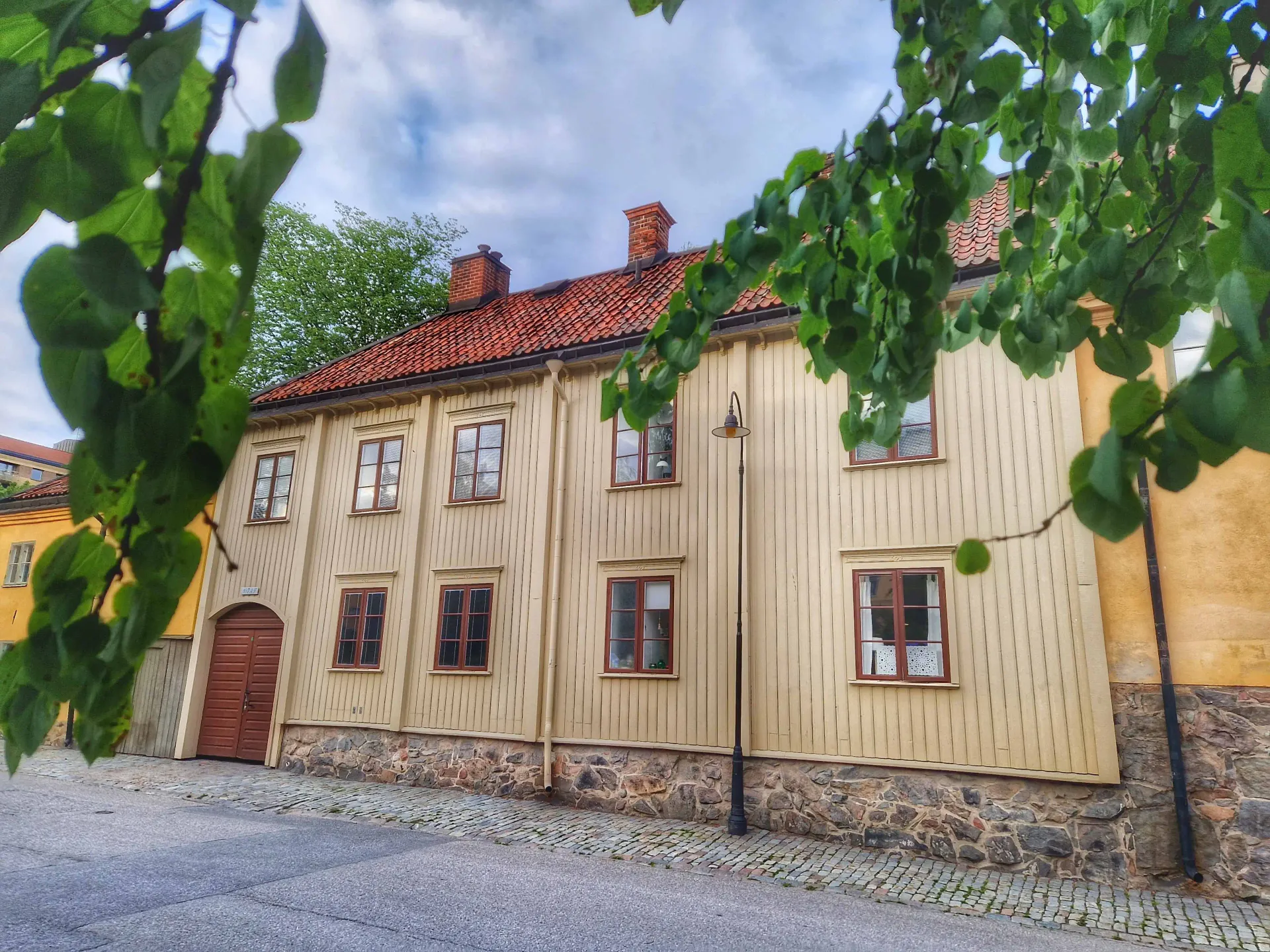 Ljust gult trähus med tegeltak vid kullerstensgata, delvis skymt av gröna grenar