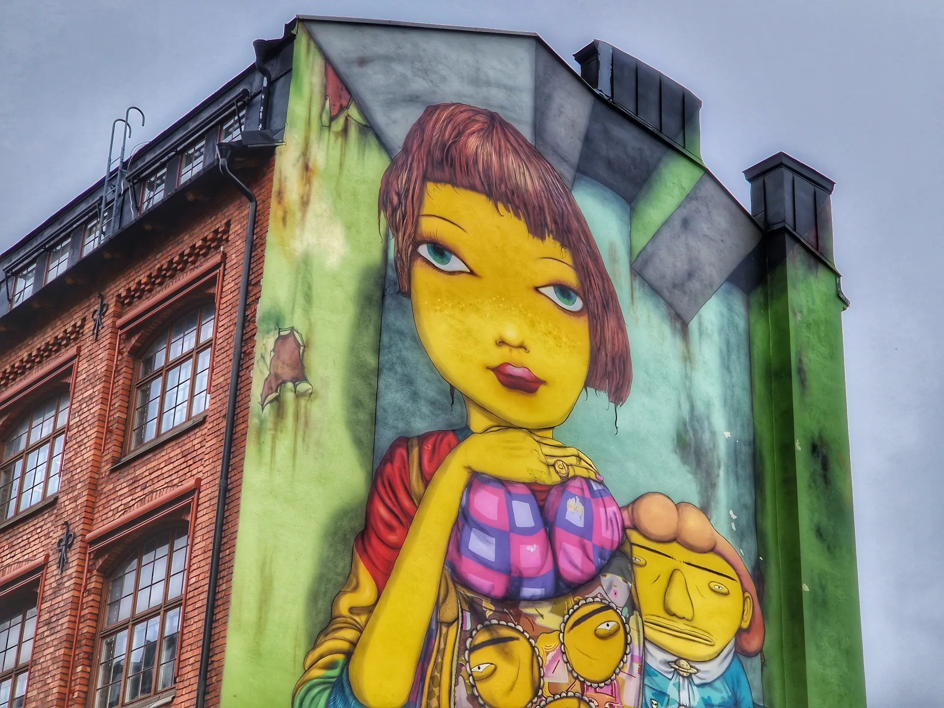 Fasadmålning med färgstark figurativ konst på husvägg bredvid rött tegelhus