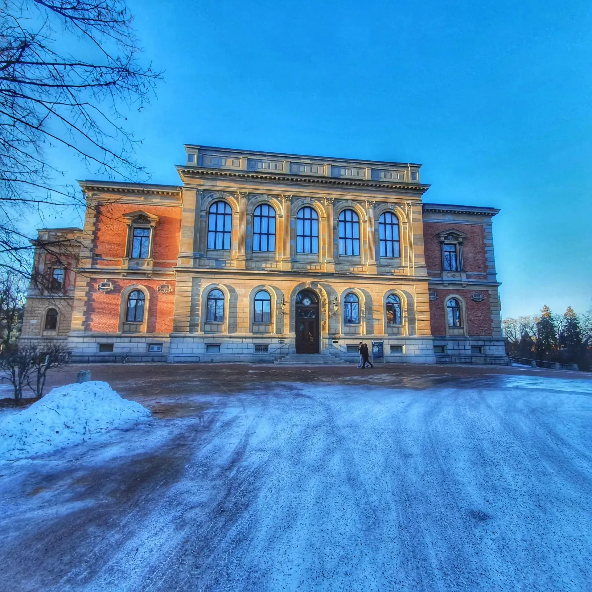 Stor byggnad i tegel och sten, två personer går in. Snö på marken
