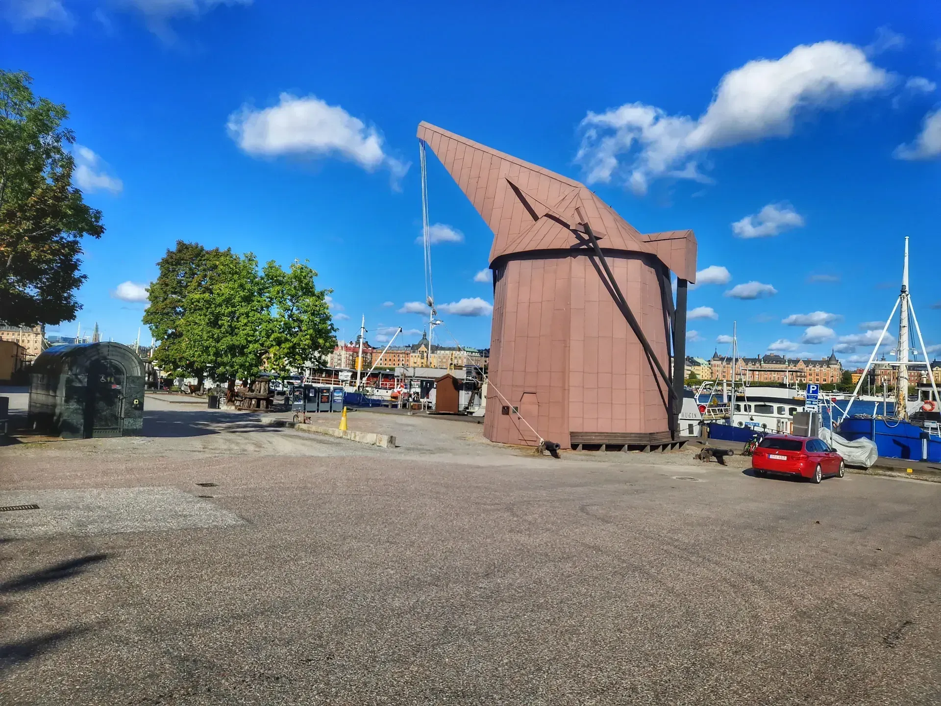 Brun kranliknande byggnad vid kaj. Röda bilar och båtar syns också. Blå himmel