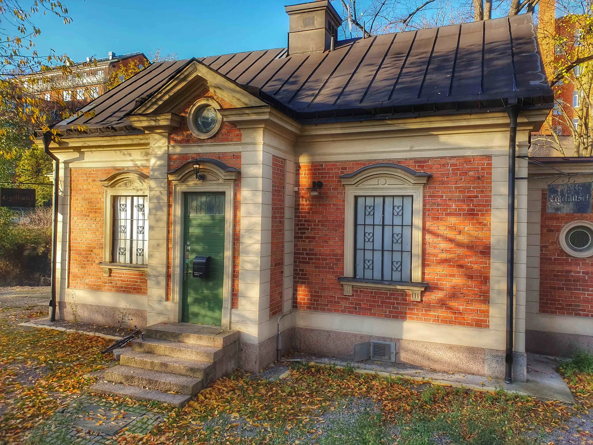 Lågt rött tegelhus med grön dörr och dekorativa fönster. Löv på marken. Skylt: "Laghuset"