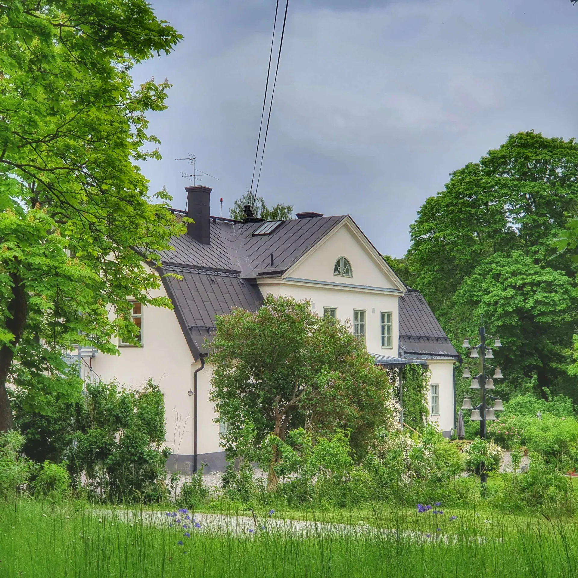 Vit byggnad med svart plåttak syns bakom träd och grönska. Ett klocktorn står till höger om huset