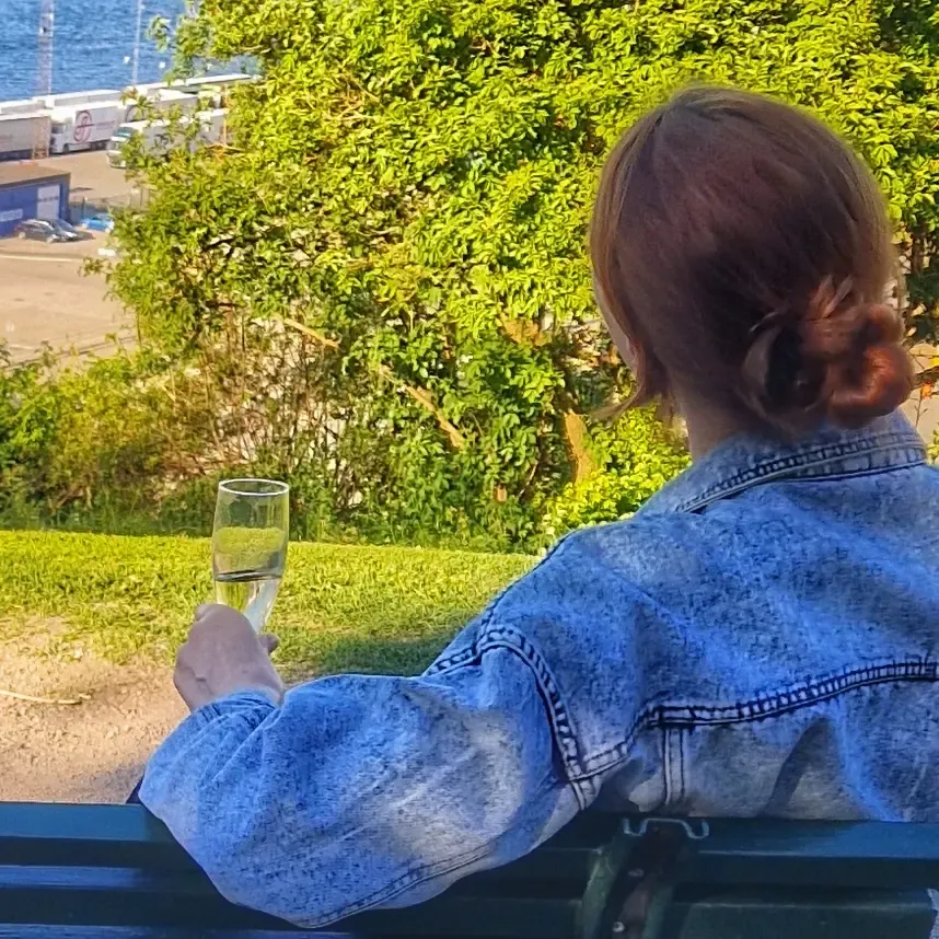 Kvinna sitter på bänk med champagne, hav och grönska i bakgrunden. Stadsvandring Södermalm