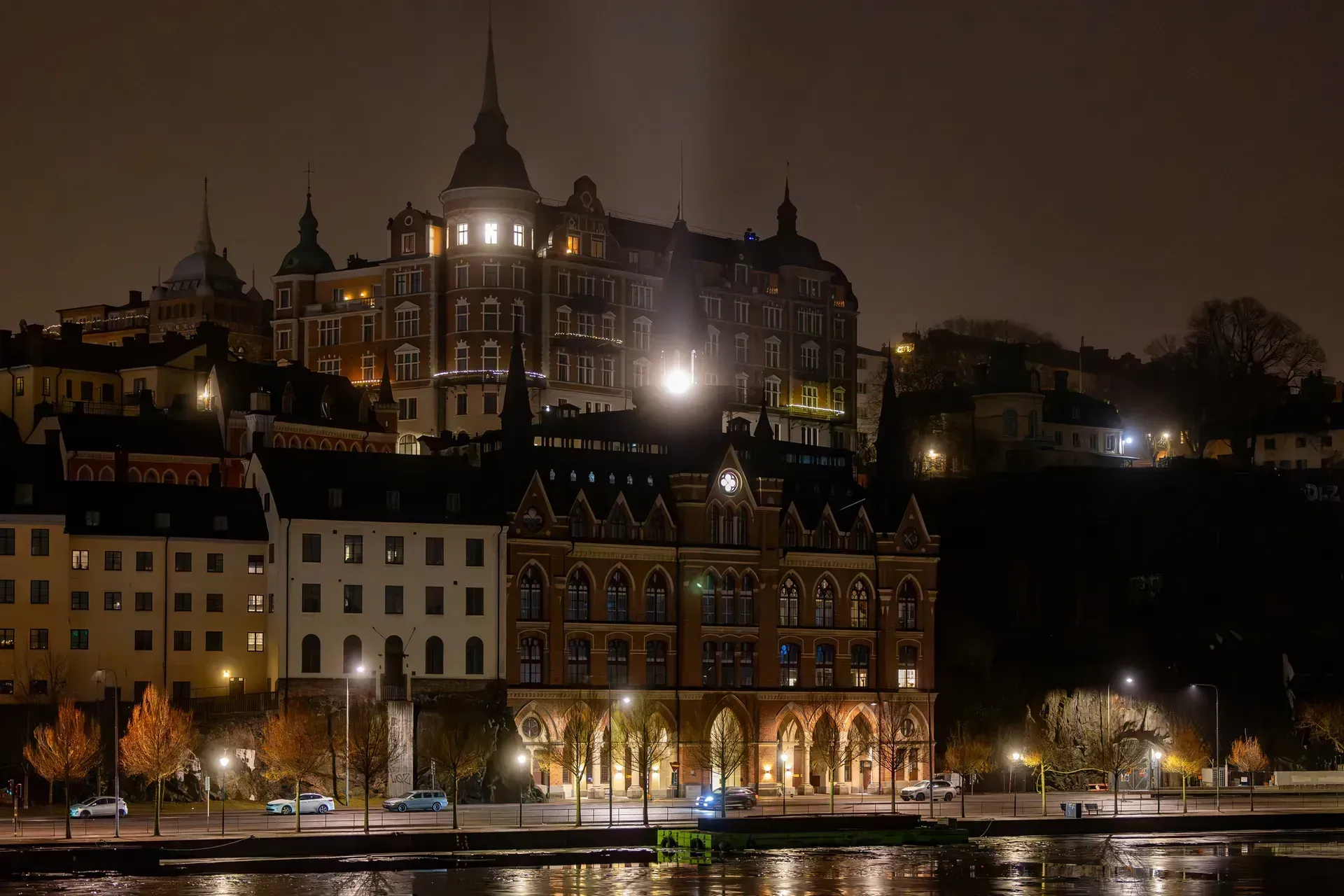 Nattvy över byggnader och kaj vid vatten. Ljus reflekteras i vattnet. Söders höjder syns i bakgrunden