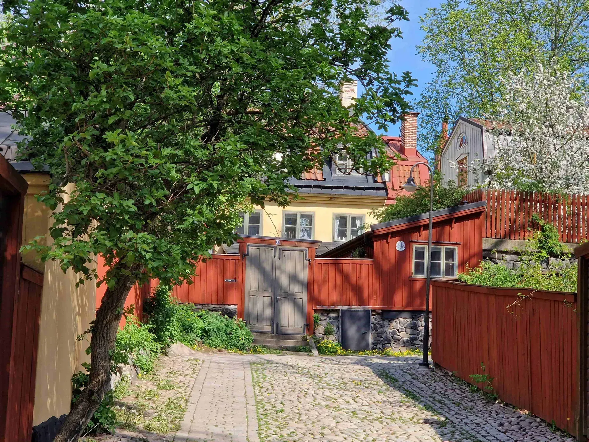Gata med kullersten, röda trästaket och trähus. Grönska. Soligt. Mariaberget Södermalm