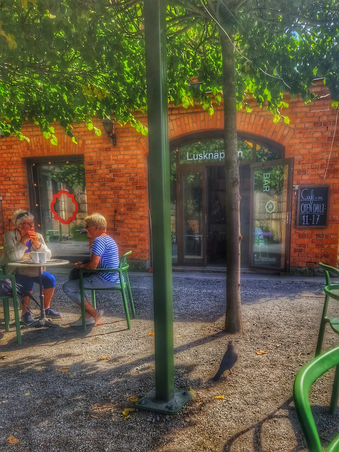 Café med två kvinnor utomhus. Tegelbyggnad, grusgång, bord, stolar och en fågel