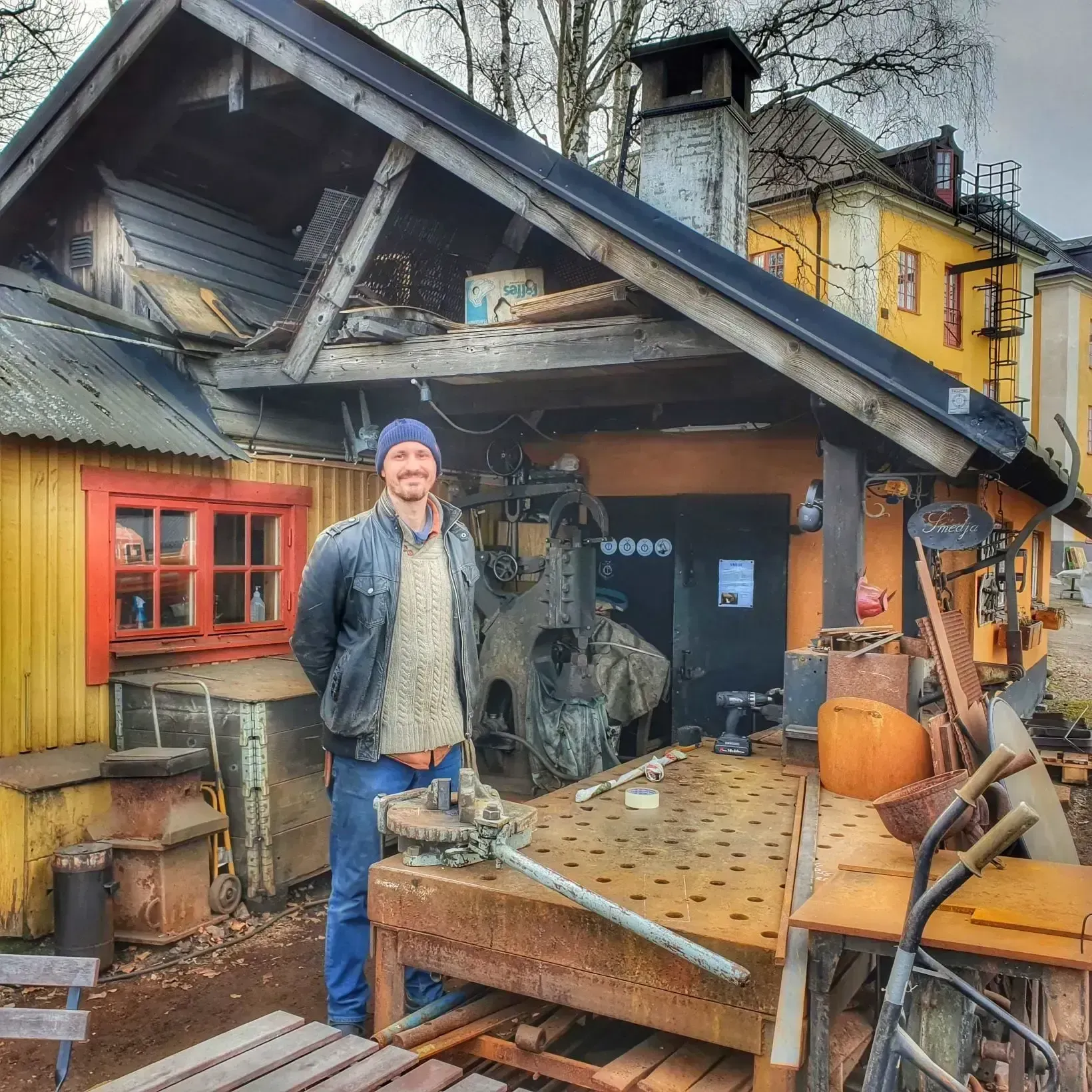 Man i blå mössa vid smedja. Röd fönsterkarm på gul byggnad. Verktyg, maskiner och material