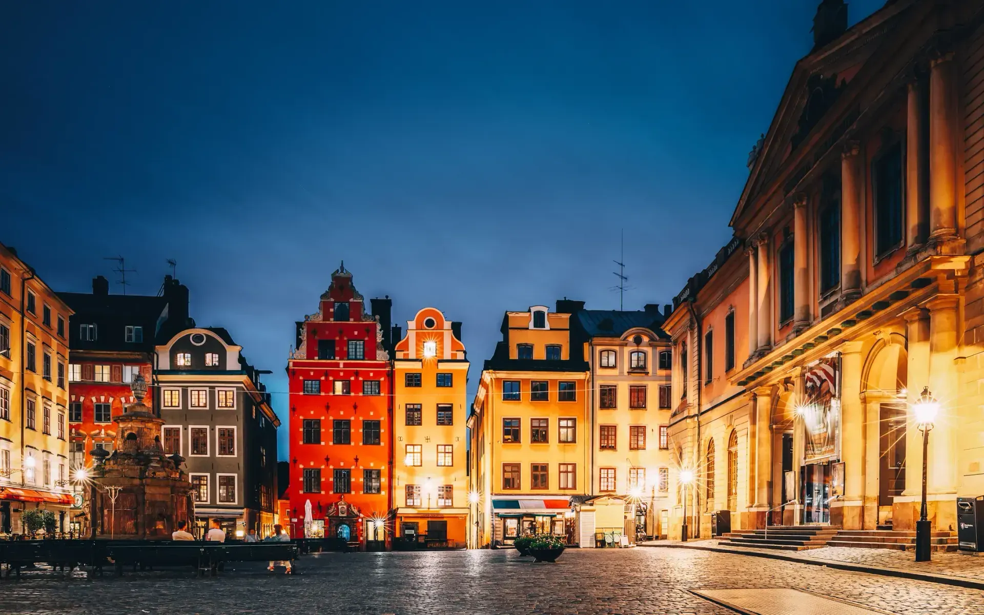 Färgglada husfasader och gatstenstorg i Gamla Stan i Stockholm under mörkblå kvällshimmel. Fontän och bänkar