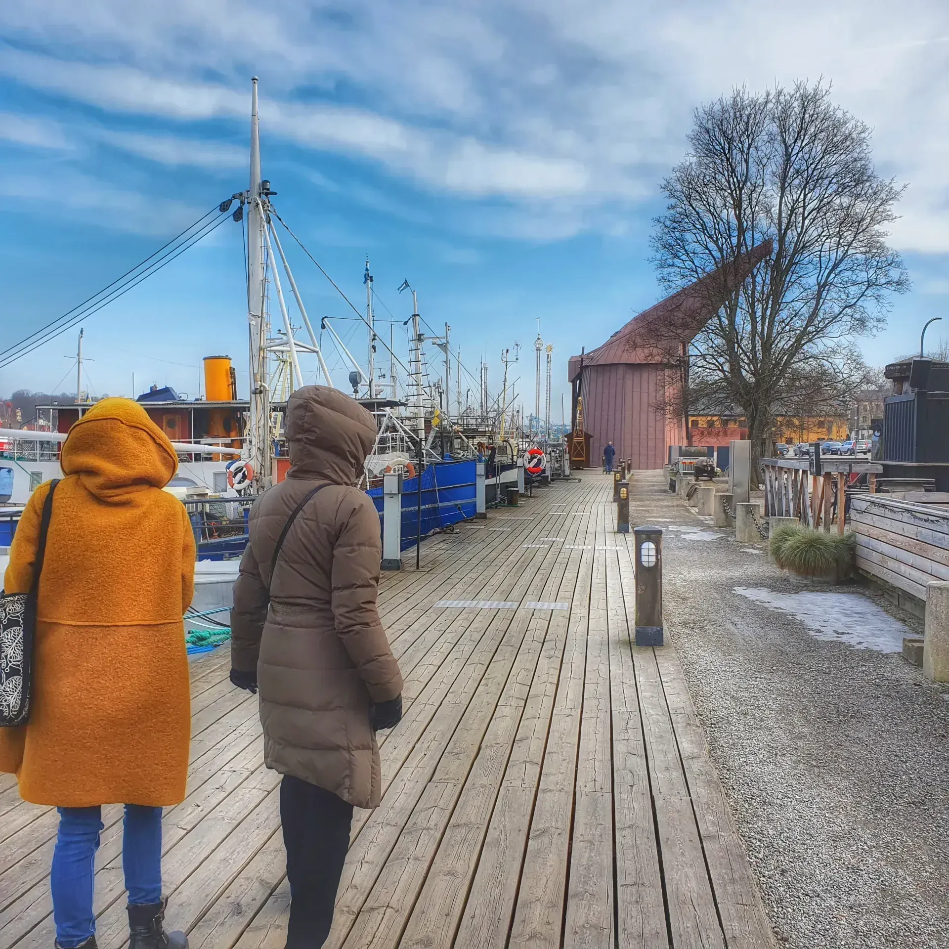 Två kvinnor promenerar på brygga. Fartyg, träd och byggnader i bakgrunden
