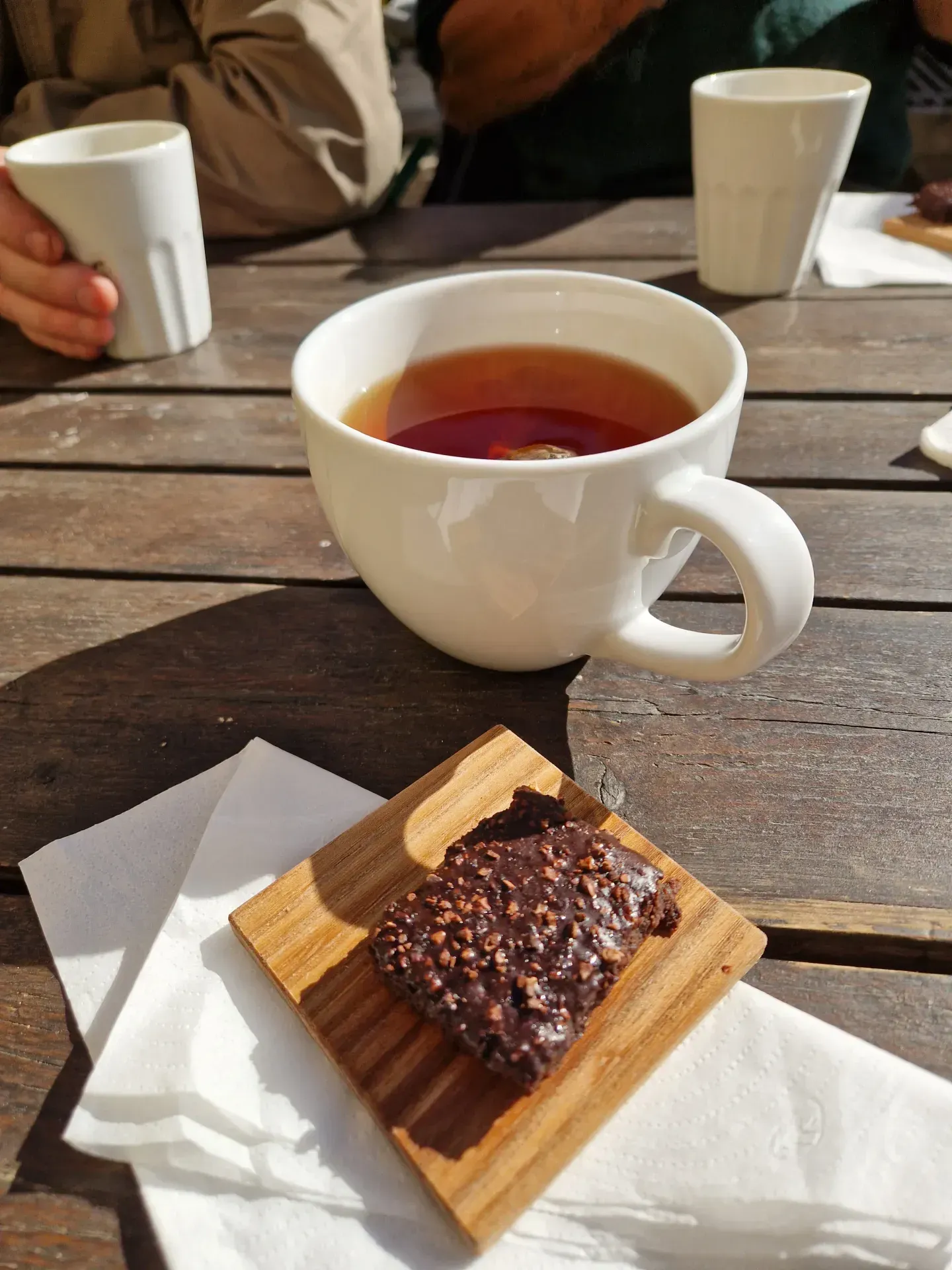 Tekopp och chokladruta på träbord. Vita servetter. Andra personer syns i bakgrunden