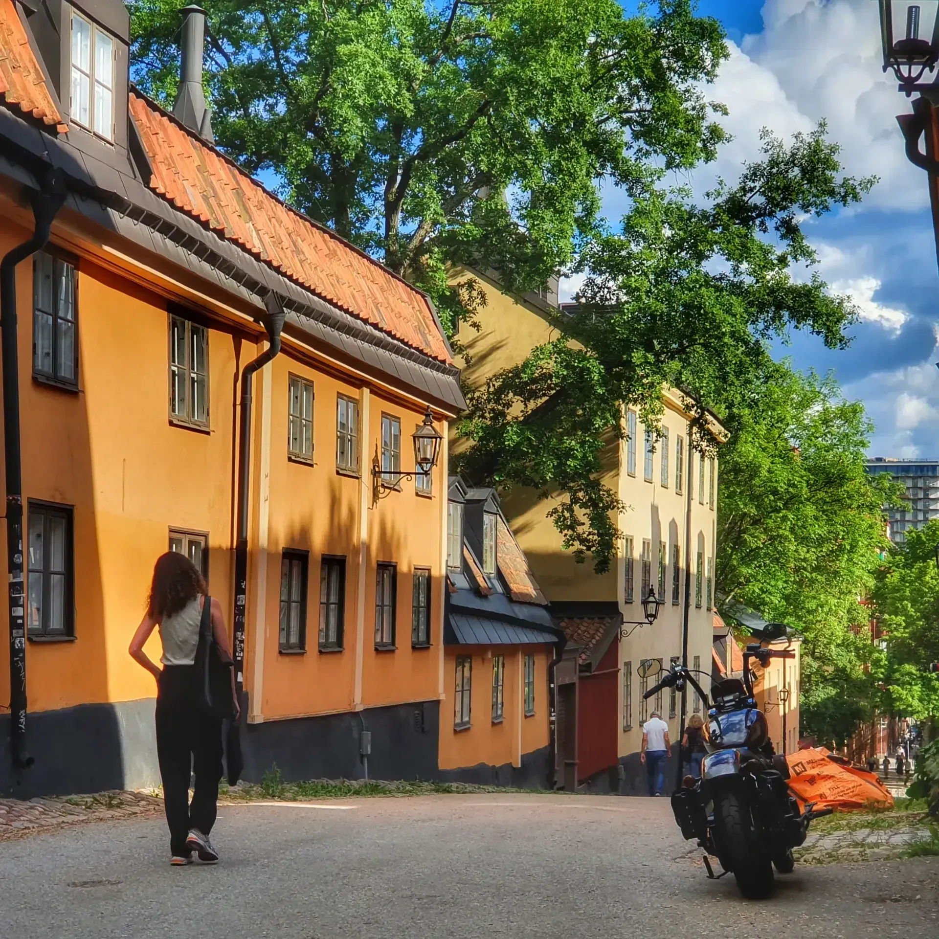 Gata i Mariaberget Södermalm med hus, träd, kvinna och parkerad motorcykel. Blå himmel med moln