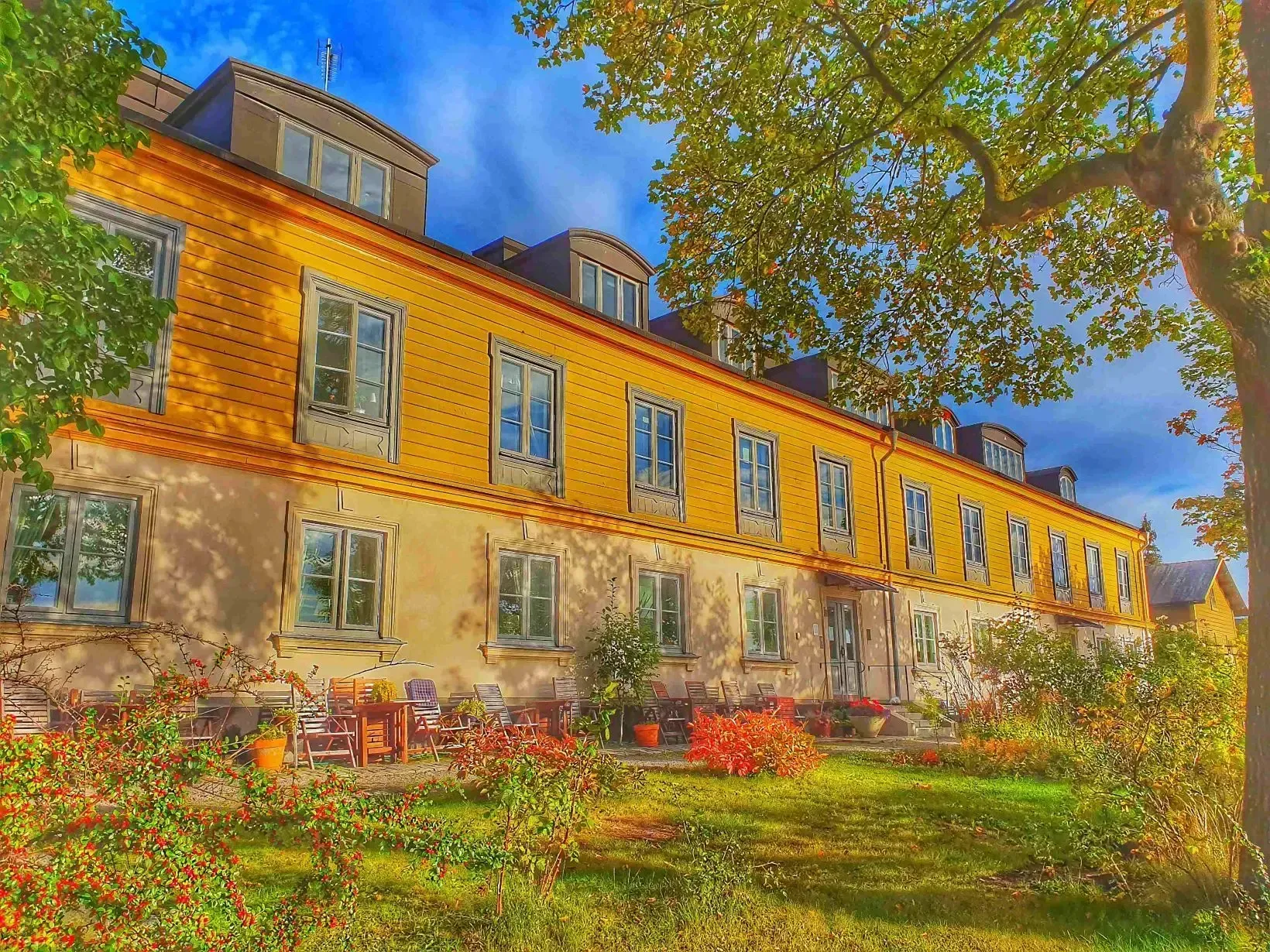 Lång, gul byggnad med många fönster. Möbler och blommor på gräsmattan framför