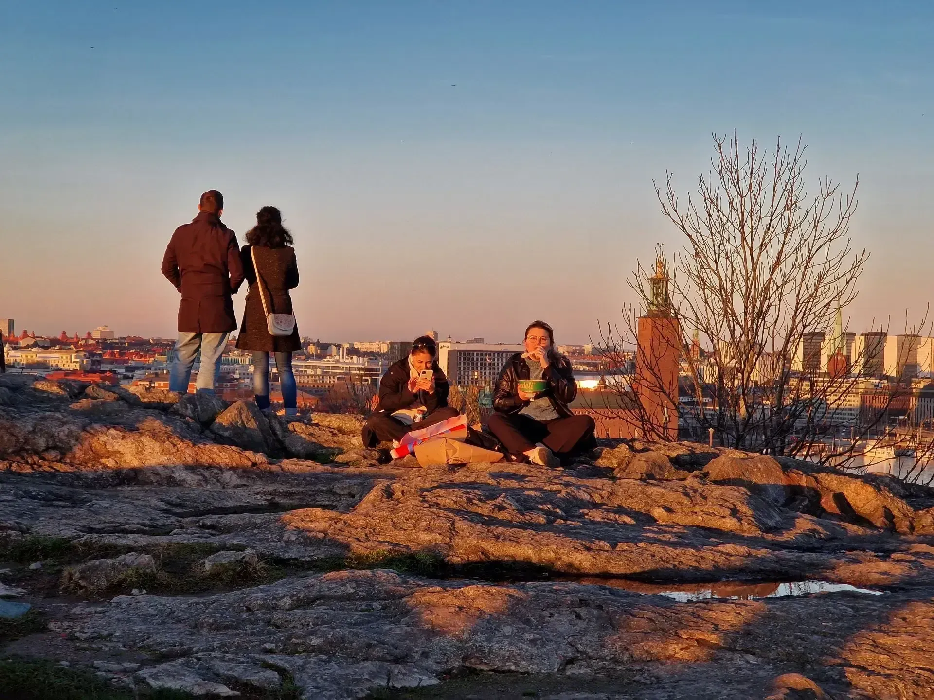 Folk på berg ser på Stockholms vy i solnedgång. Två sitter och äter. Stenigt underlag
