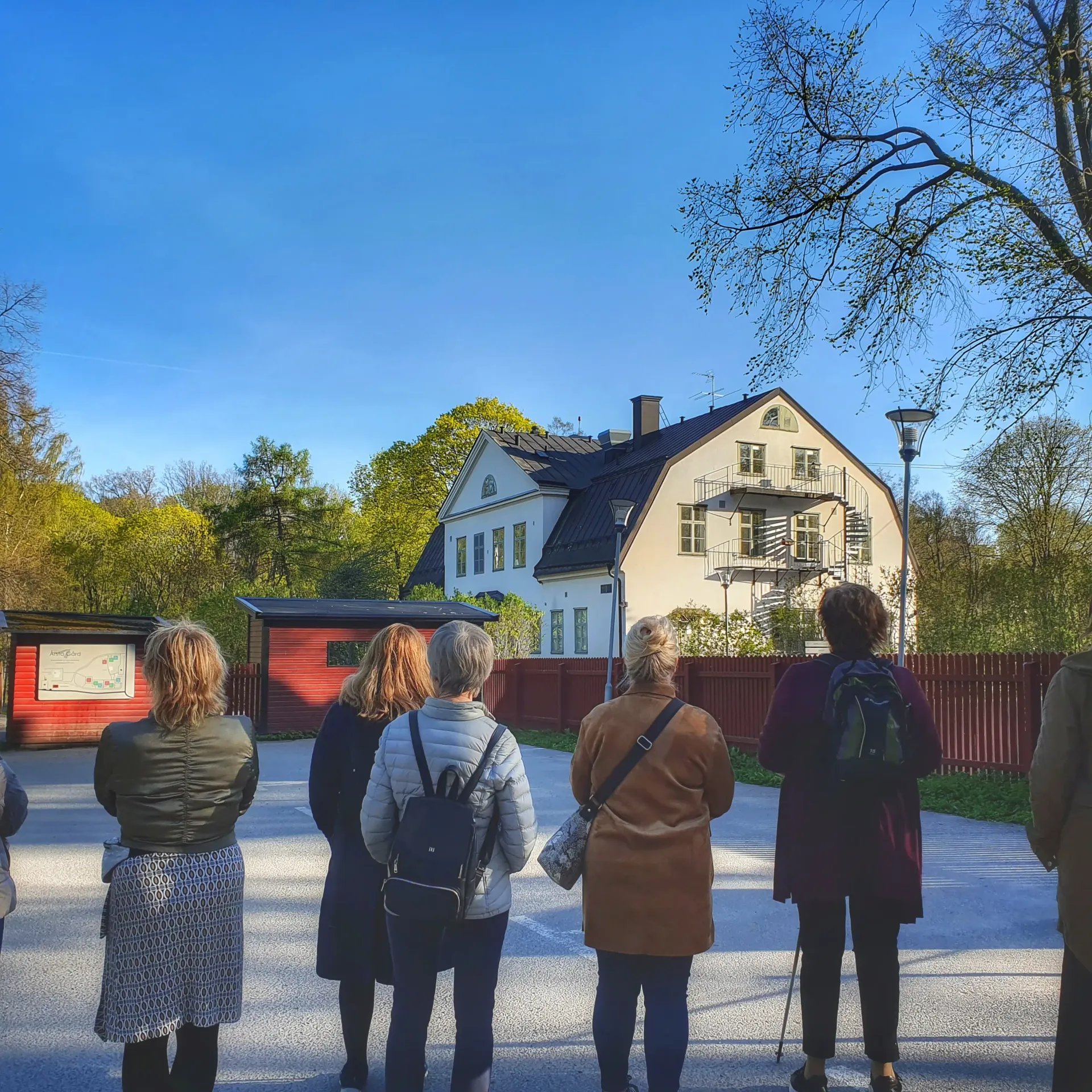 Kvinnor står framför ett hus med spiraltrappa utomhus. Röda byggnader och staket syns också