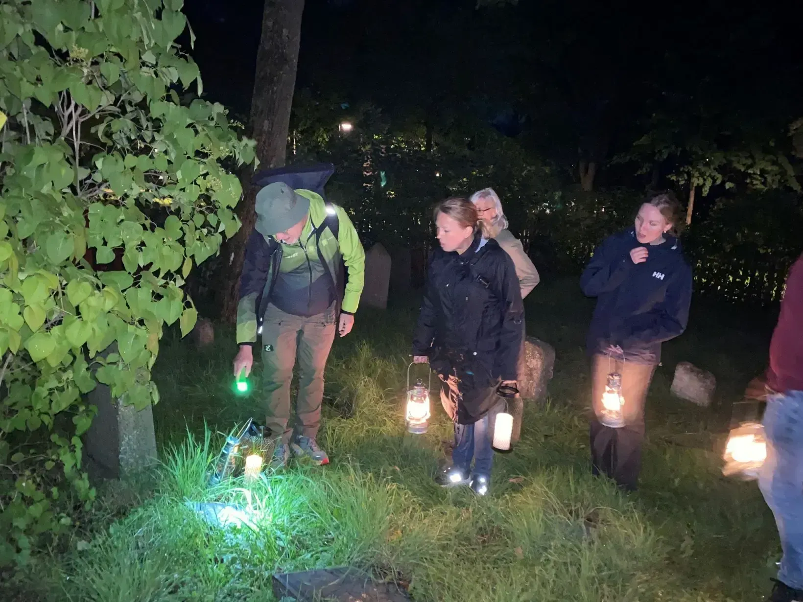 Grupp människor på en kyrkogård i mörker, lyser med ficklampa och lyktor