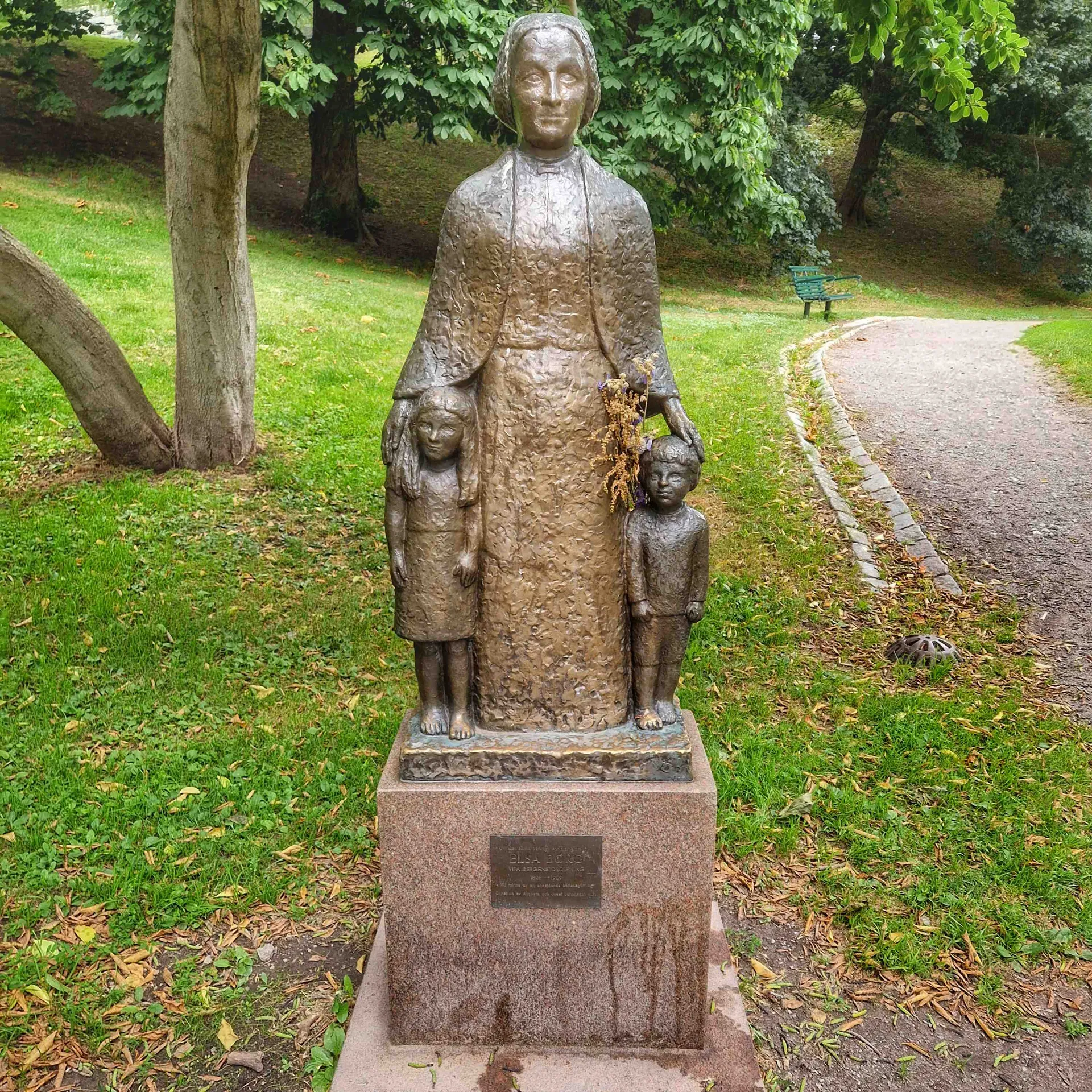 Staty av kvinna med två barn, stående på sockel i park. Gräs och träd i bakgrunden