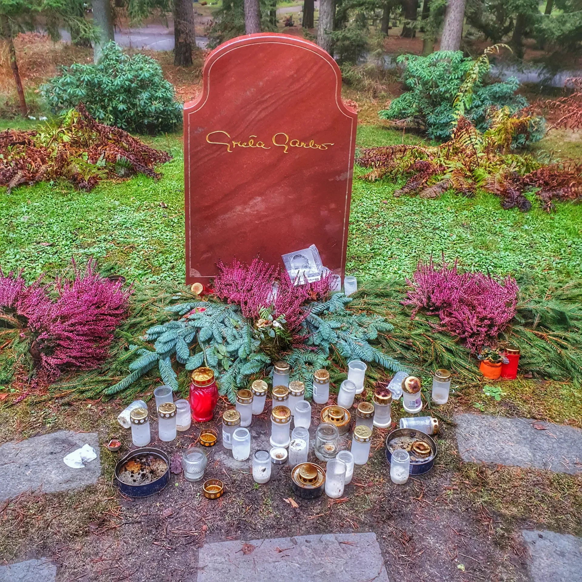 Gravsten med texten "Greta Garbo". Blommor, ljus och lyktor vid stenen. Skogskyrkogården