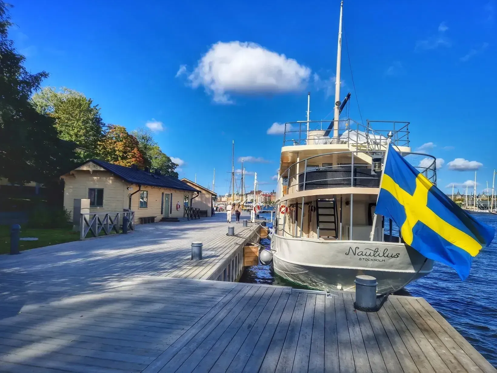 Båt med svensk flagga vid brygga. Trähus på kajen. Blå himmel med moln