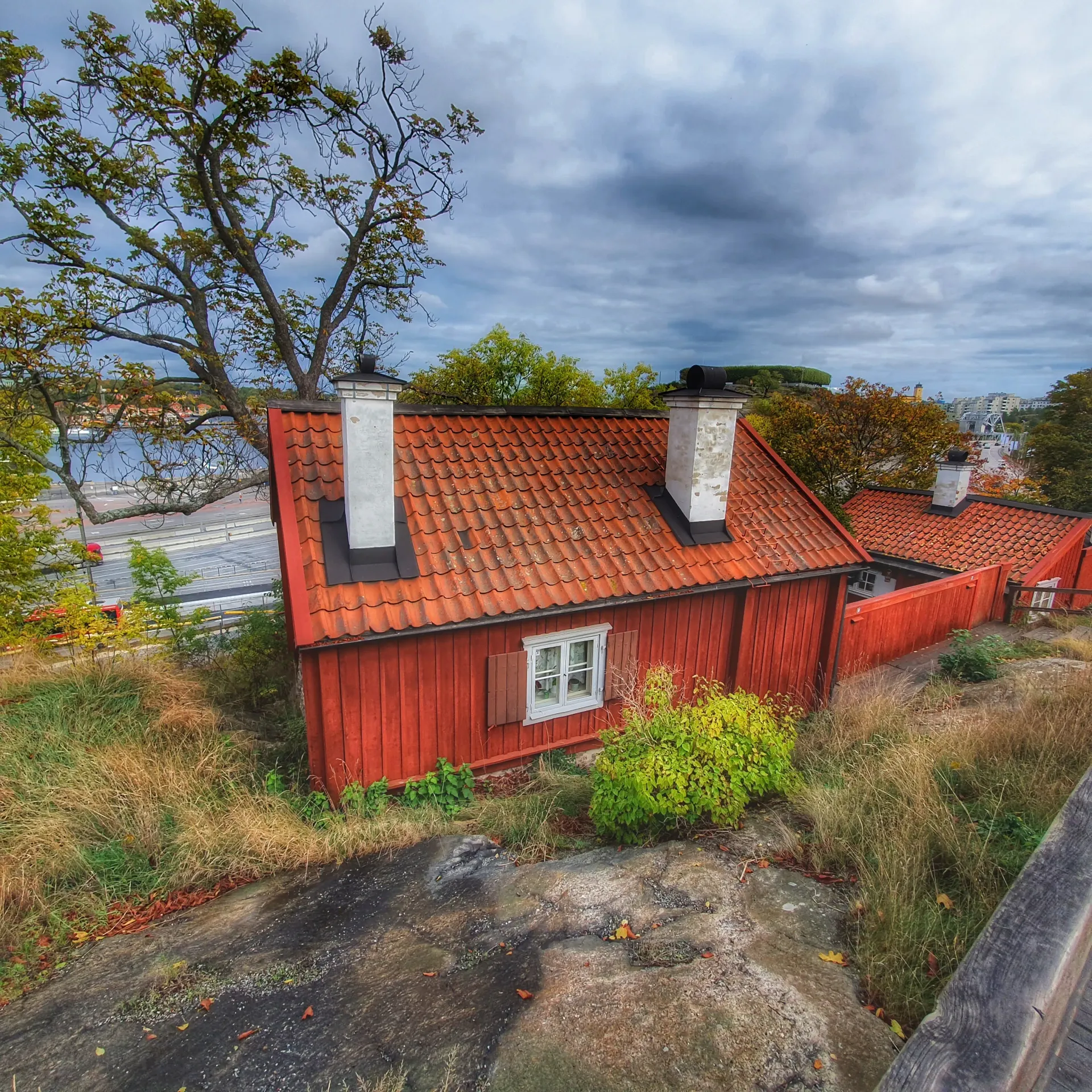 Rött trähus med tegeltak och vita skorstenar på klippig tomt. Lövträd och gräs. Molnig himmel. "Smaka på historien"