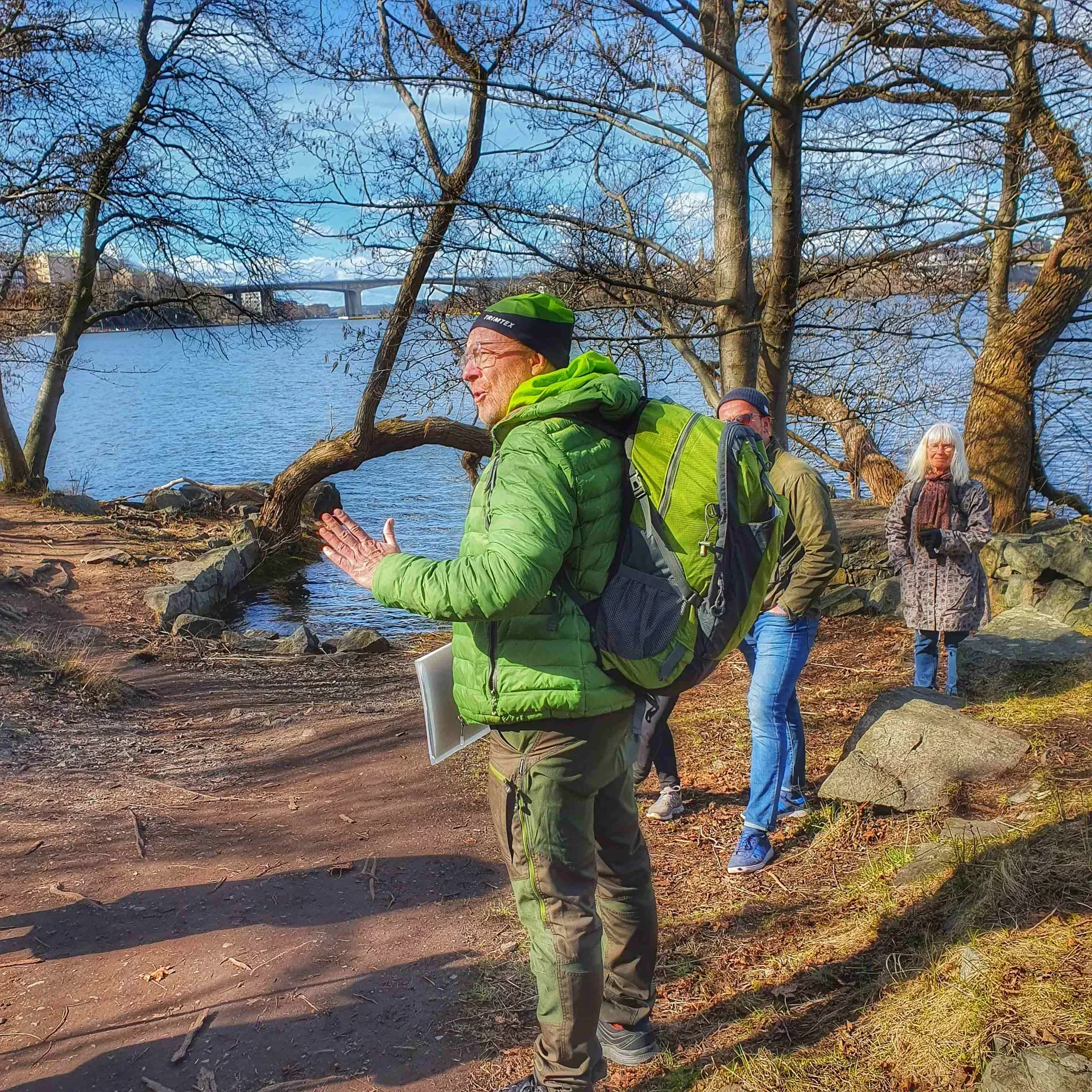 Grupp människor vid vattnet. Man i grön jacka med ryggsäck gestikulerar. Träd och bro i bakgrunden