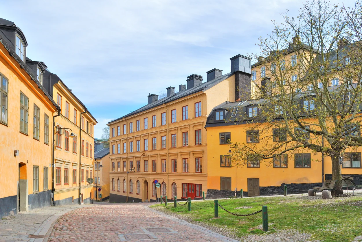 Stenlagd gata mellan gula hus, en vanlig syn vid stadsvandring på Mariaberget. Till höger träd och bänk på gräs